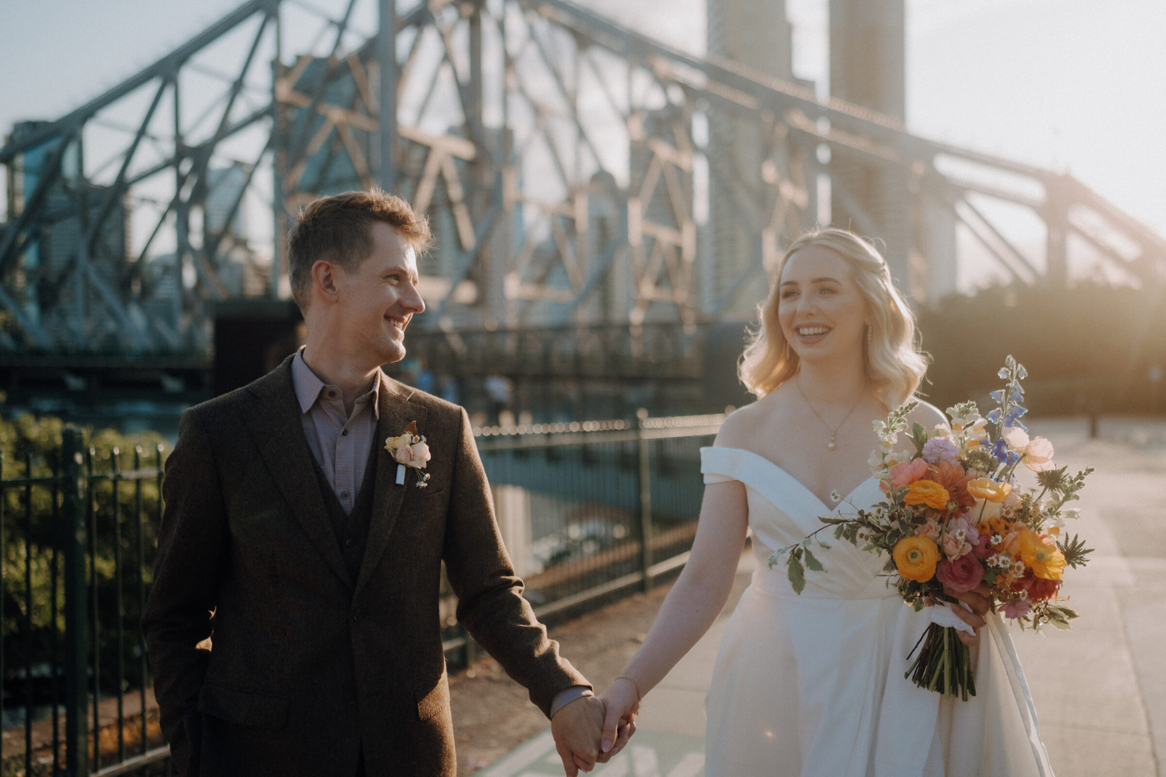 A couple in wedding attire holds hands and smiles while walking near a large bridge in the background. The bride holds a bouquet of colorful flowers.