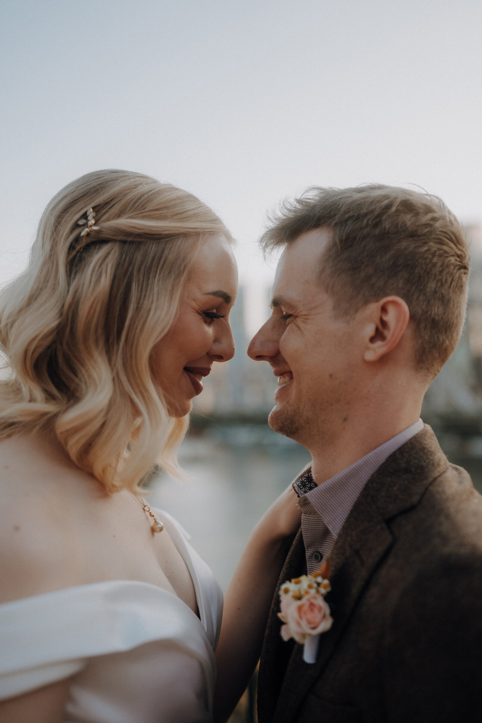 A couple stands close, smiling at each other. The woman wears a white dress, and the man wears a suit with a boutonniere. The background is blurred.