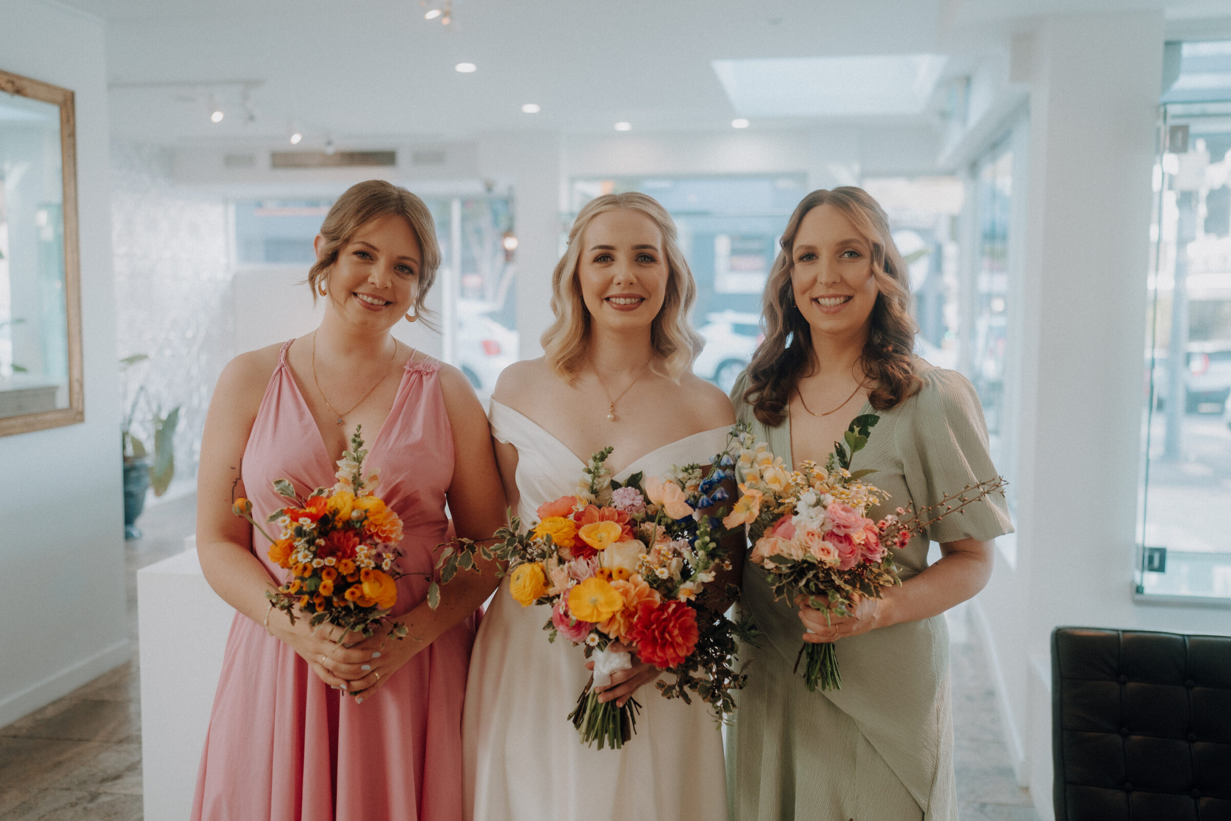 Three women in formal attire holding colorful bouquets stand indoors, smiling at the camera.