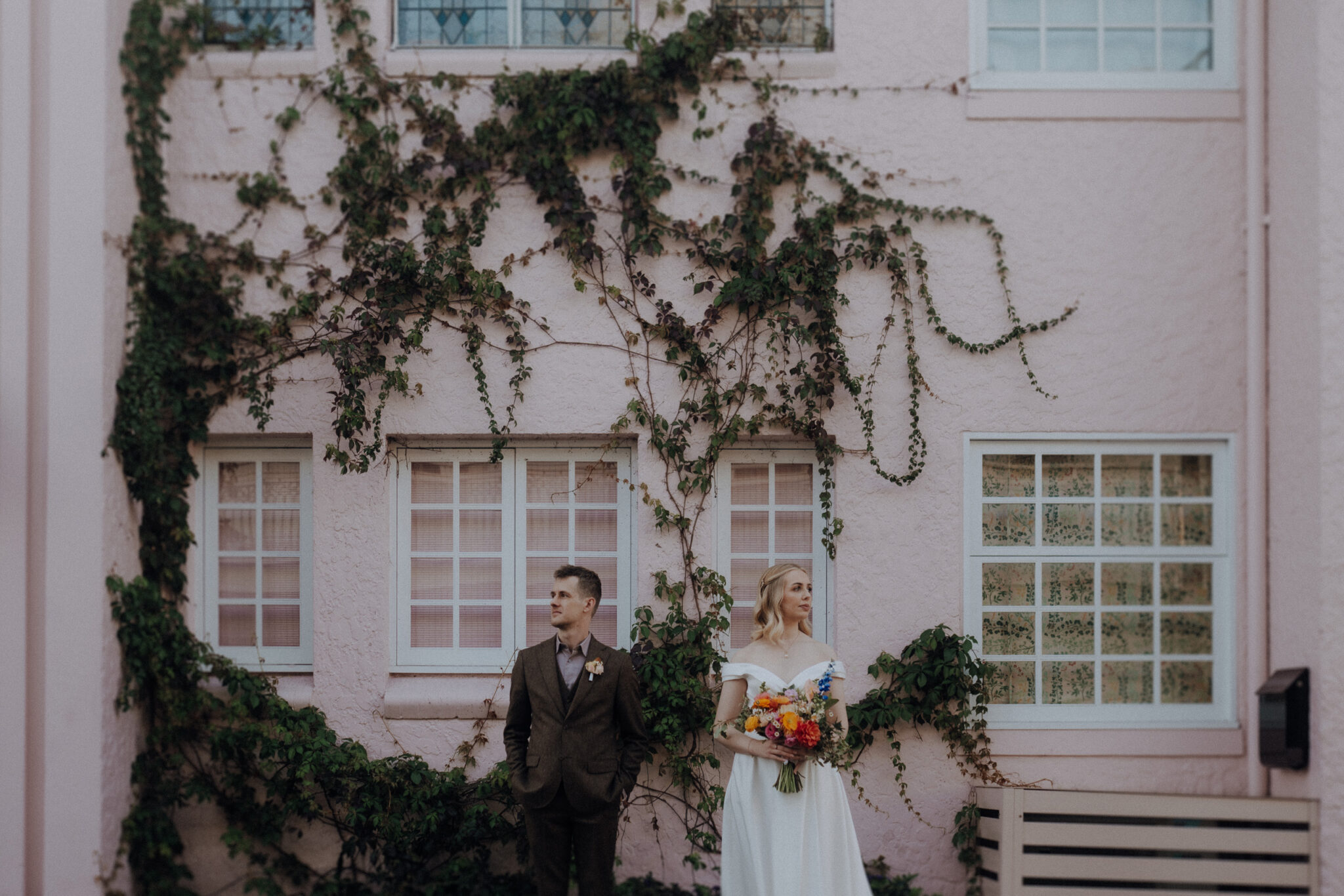 A couple stands apart in front of a pink building with vines. The woman holds a bouquet.