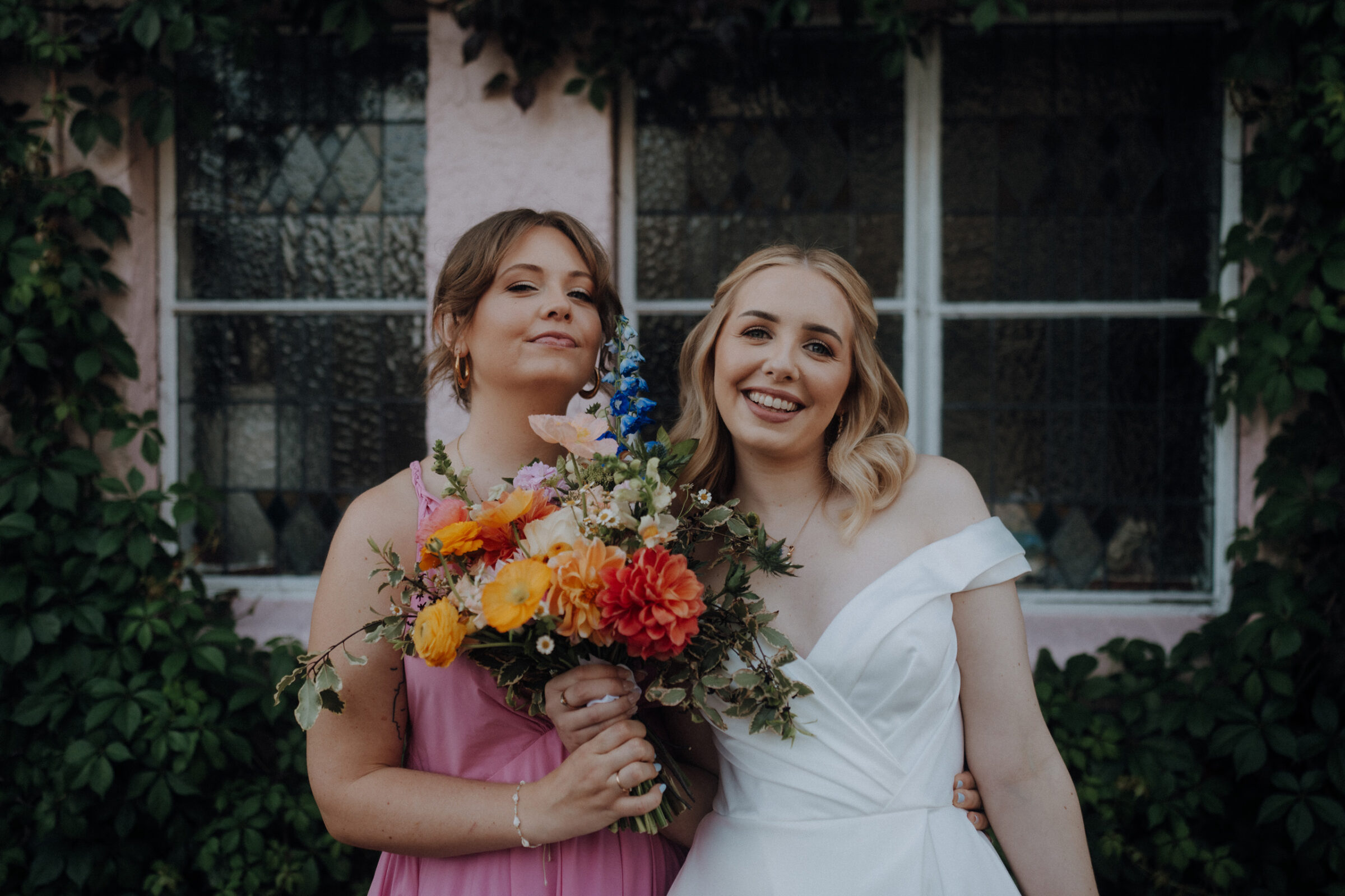 Two women stand smiling; one in a white dress and the other in pink, holding a colorful bouquet, against a background of ivy-covered windows.