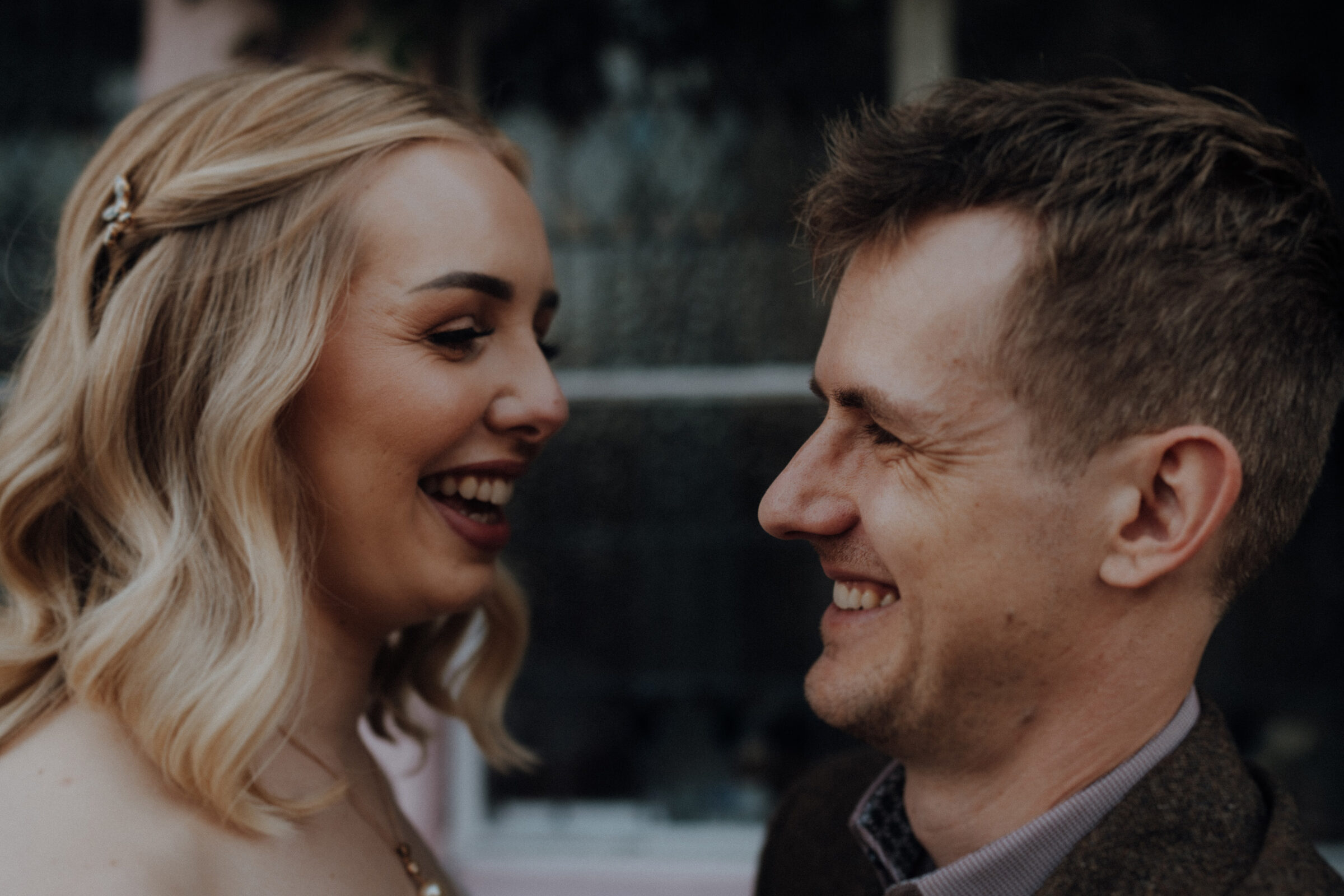 A woman and a man stand facing each other, smiling warmly. The background is softly blurred.