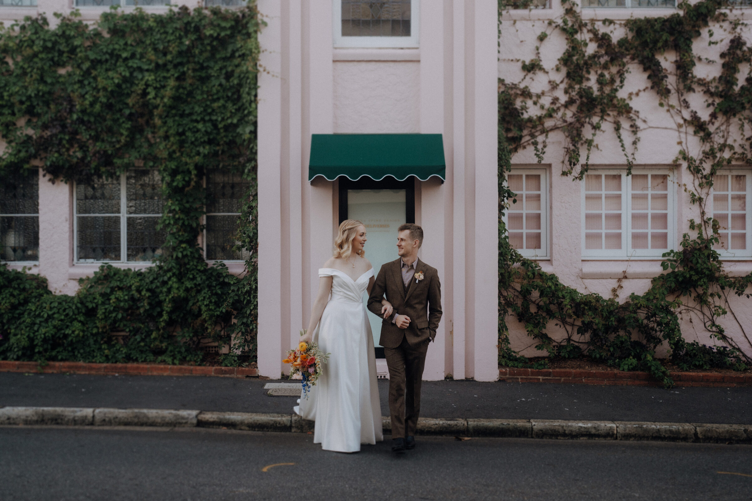 A couple in wedding attire walks hand in hand in front of a pink building with green ivy and a green awning.