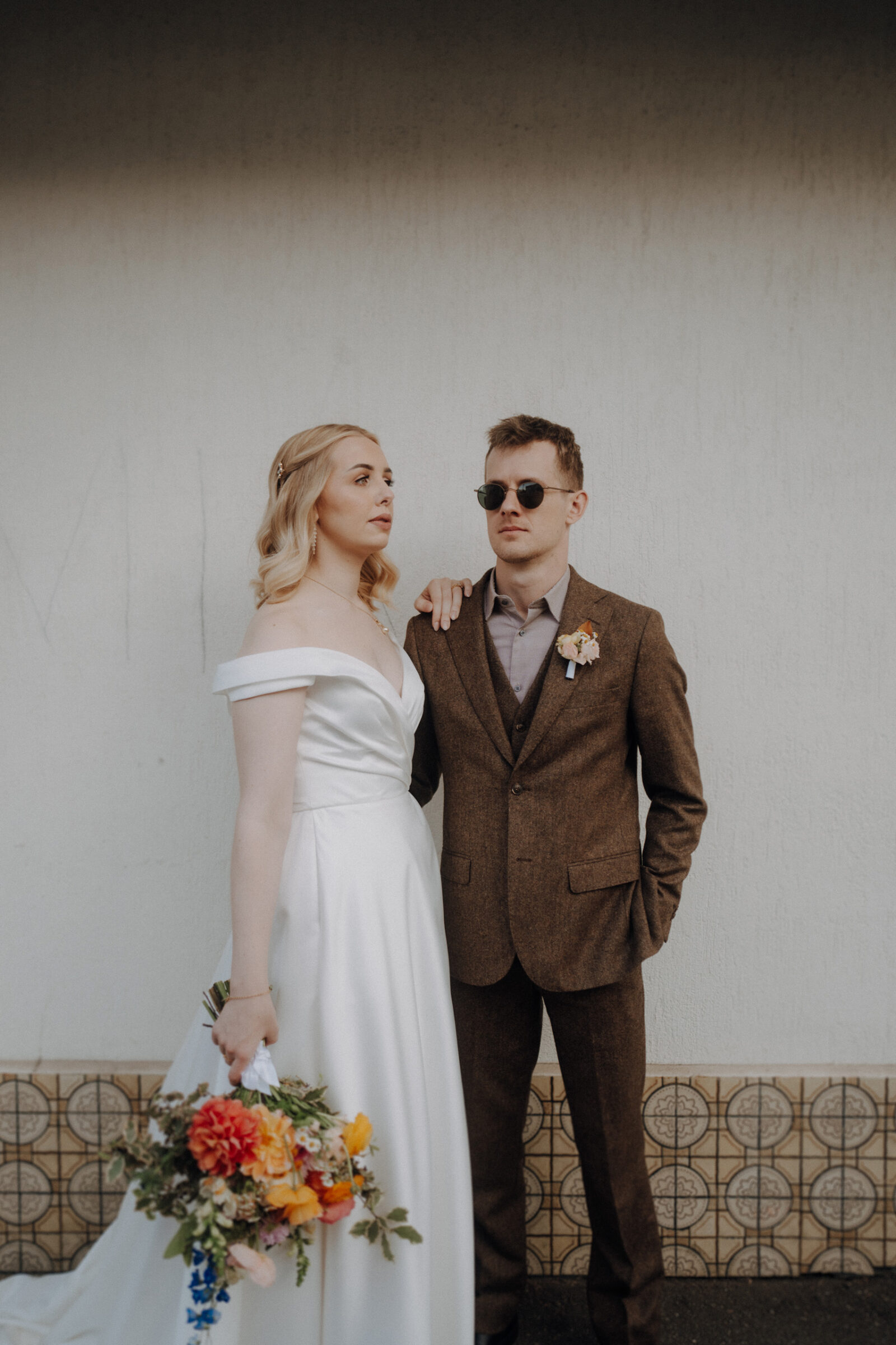 Bride in a white gown holds a colorful bouquet, standing next to a groom in a brown suit and sunglasses, both against a plain wall.