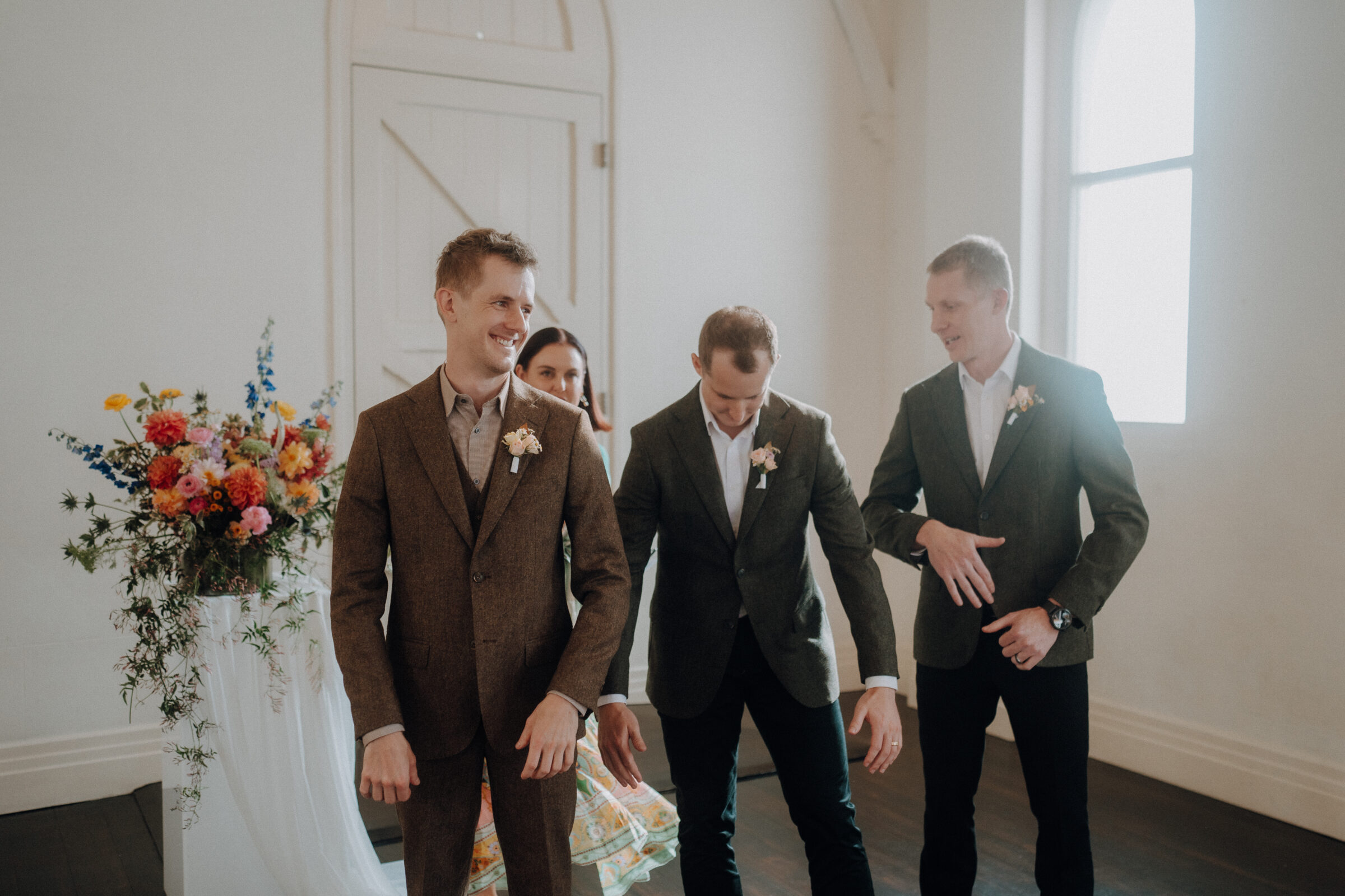 Three men in suits stand in a bright room with floral decorations, appearing to adjust their attire.