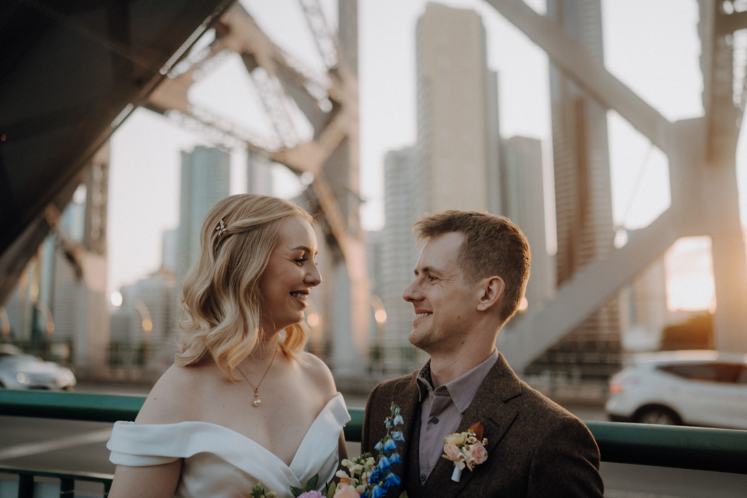 A couple in formal attire smiles at each other on a city bridge at sunset, with tall buildings in the background.