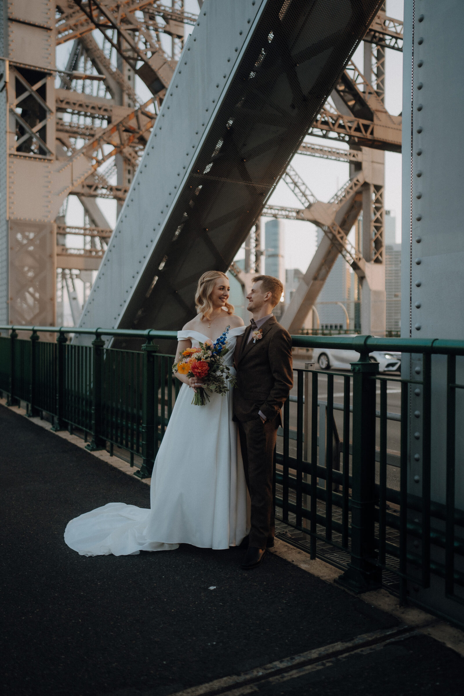 A bride and groom stand together on a bridge, with metal beams and a cityscape in the background. The bride holds a colorful bouquet.