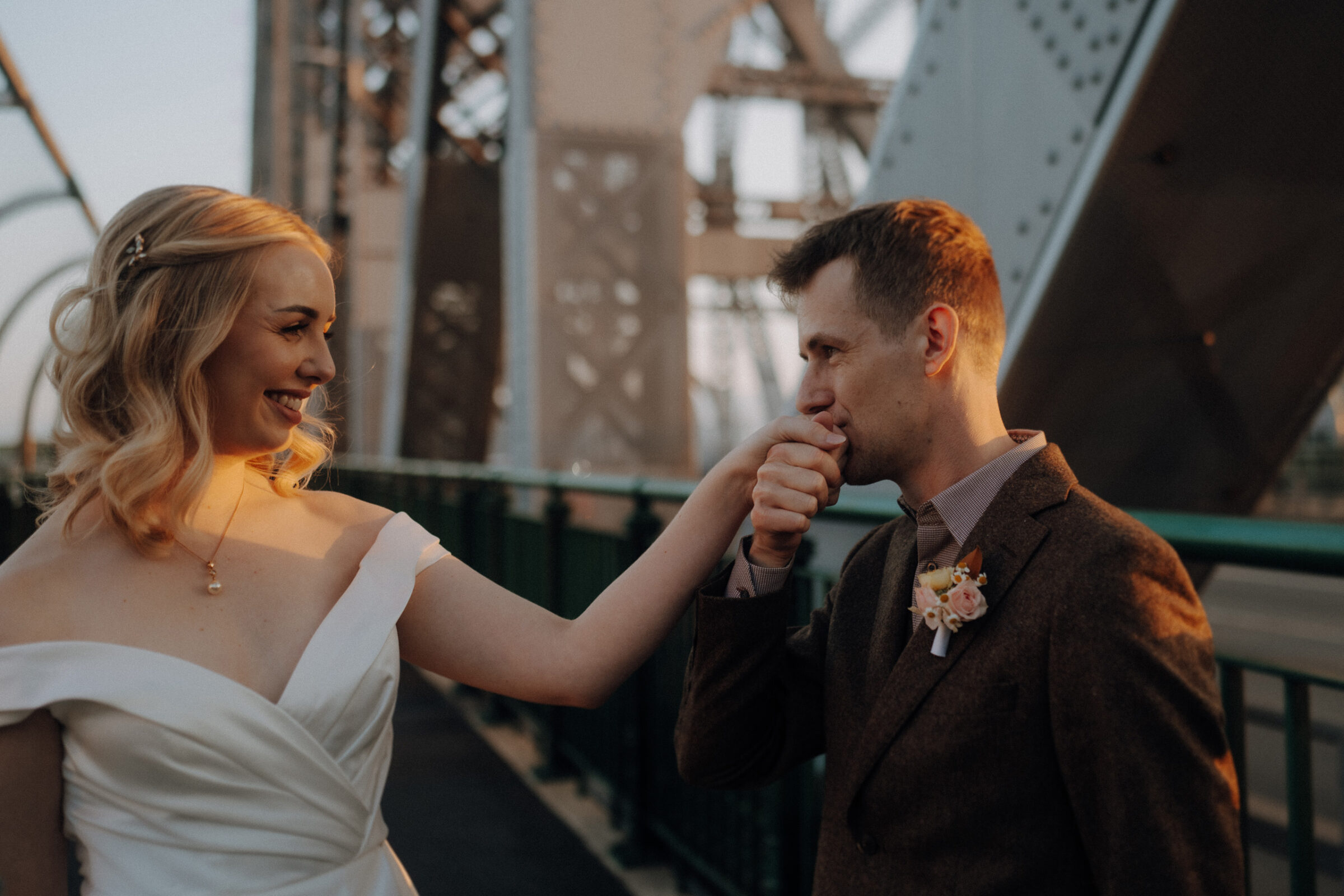 A man kisses a woman's hand on a bridge while they both smile at each other. The woman is wearing a white dress, and the man is in a suit with a boutonniere.