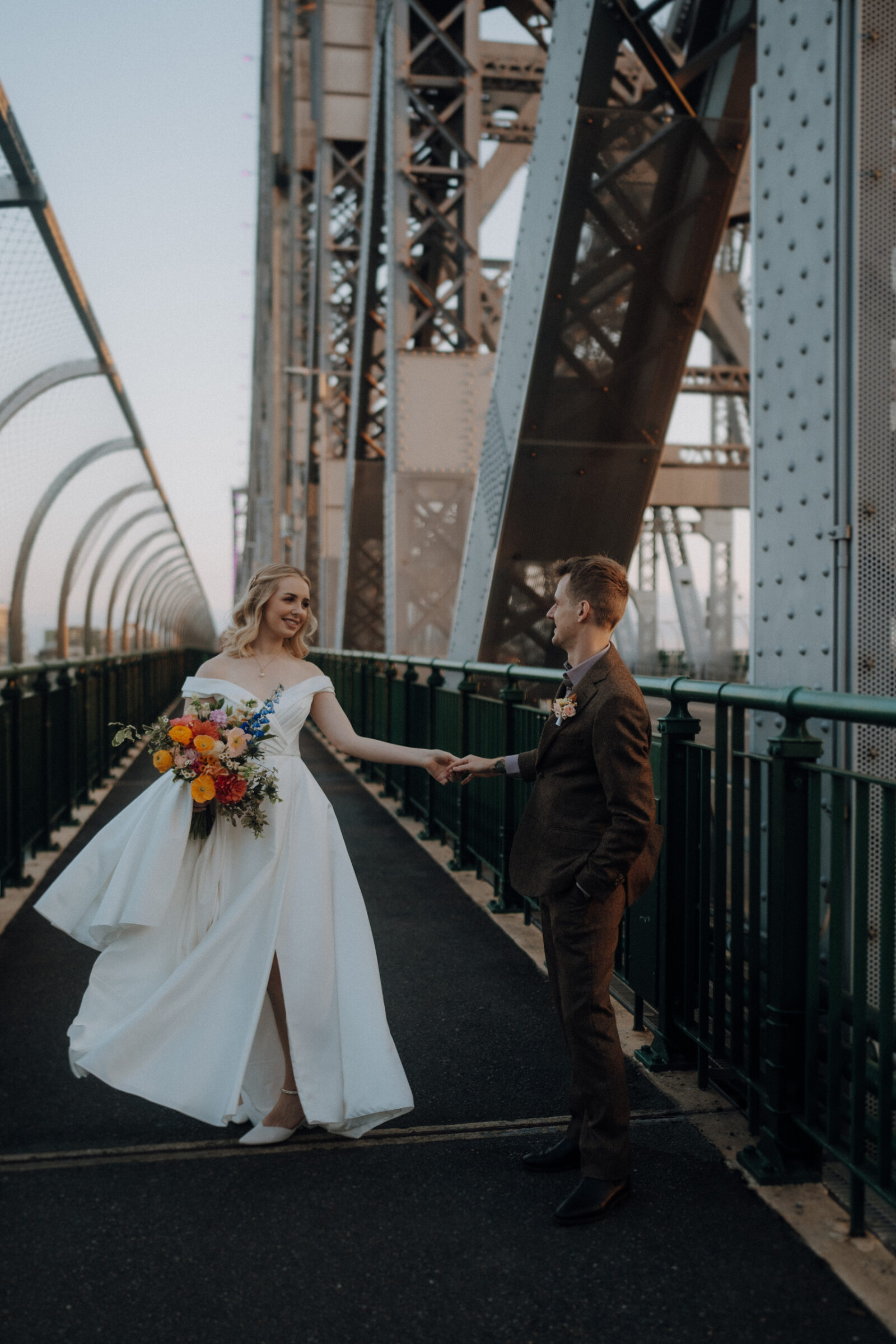 A bride and groom hold hands on a metal bridge. The bride is in a flowing white dress with a bouquet, and the groom is in a dark suit. The background shows the bridge’s steel structure.