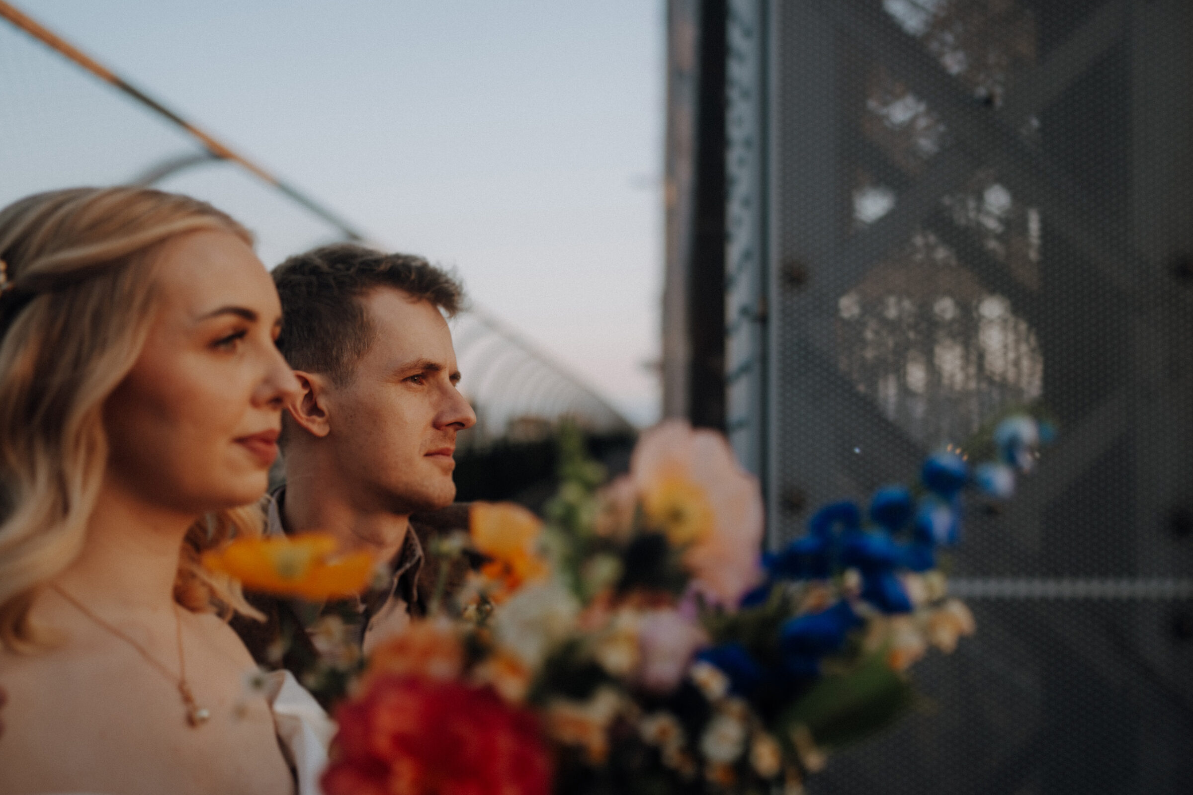 A woman and man stand side by side outdoors, holding a colorful bouquet, with a fence in the background.