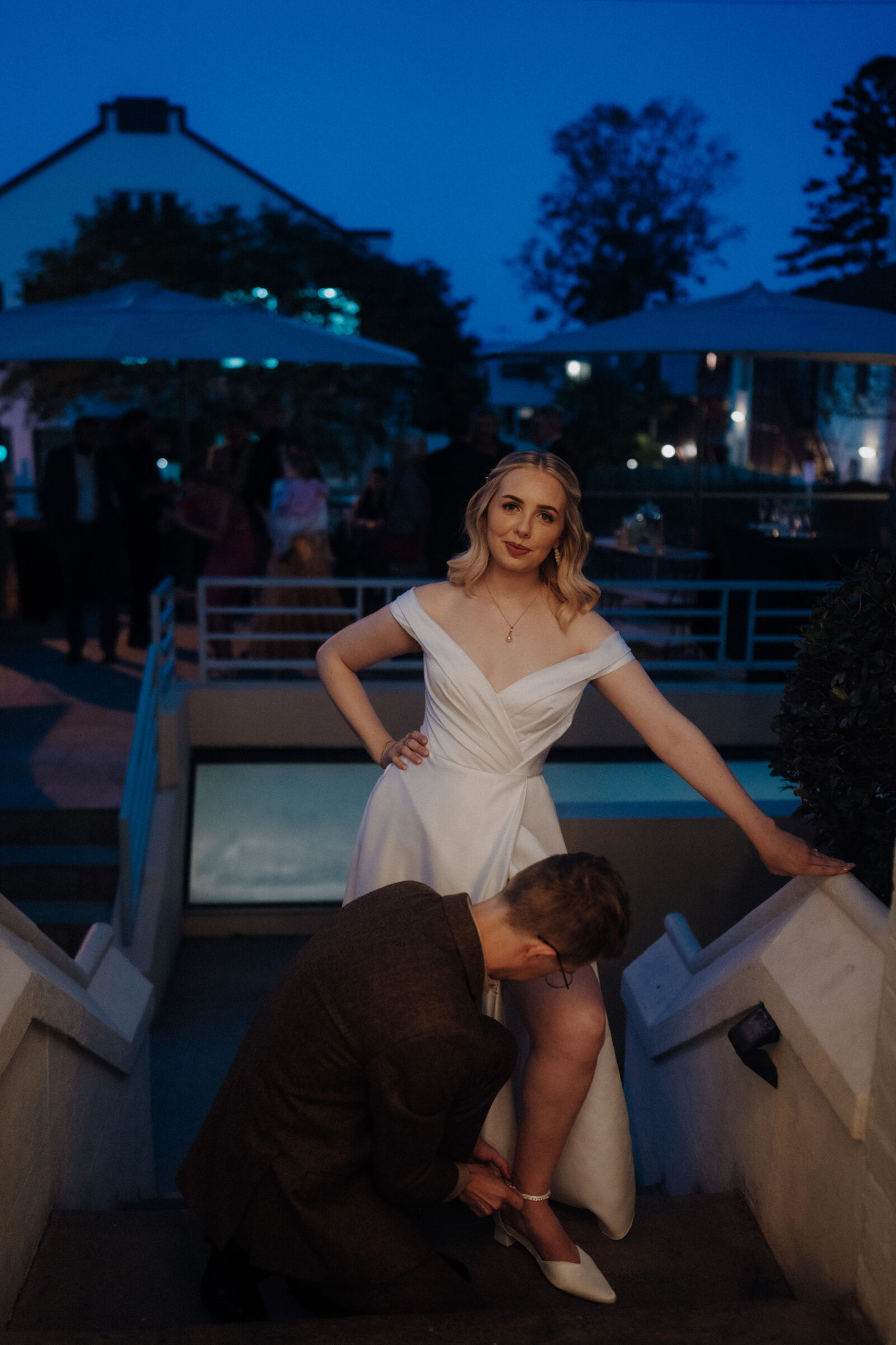 A woman in a white dress stands on stairs while a man kneels to adjust her shoe, with an evening gathering in the background.