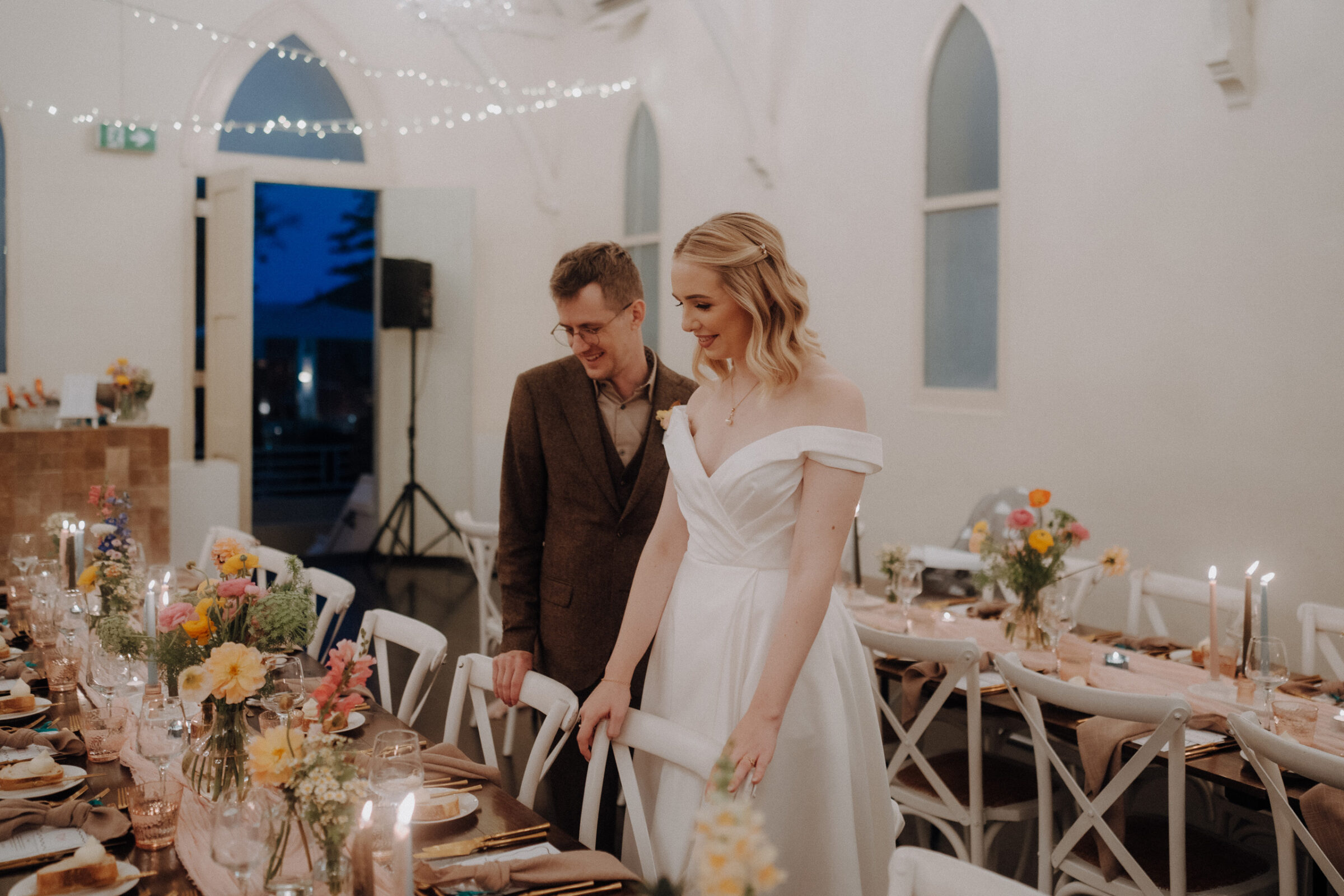 A couple stands in a softly lit room decorated for a wedding, with a long table set with flowers, candles, and place settings.