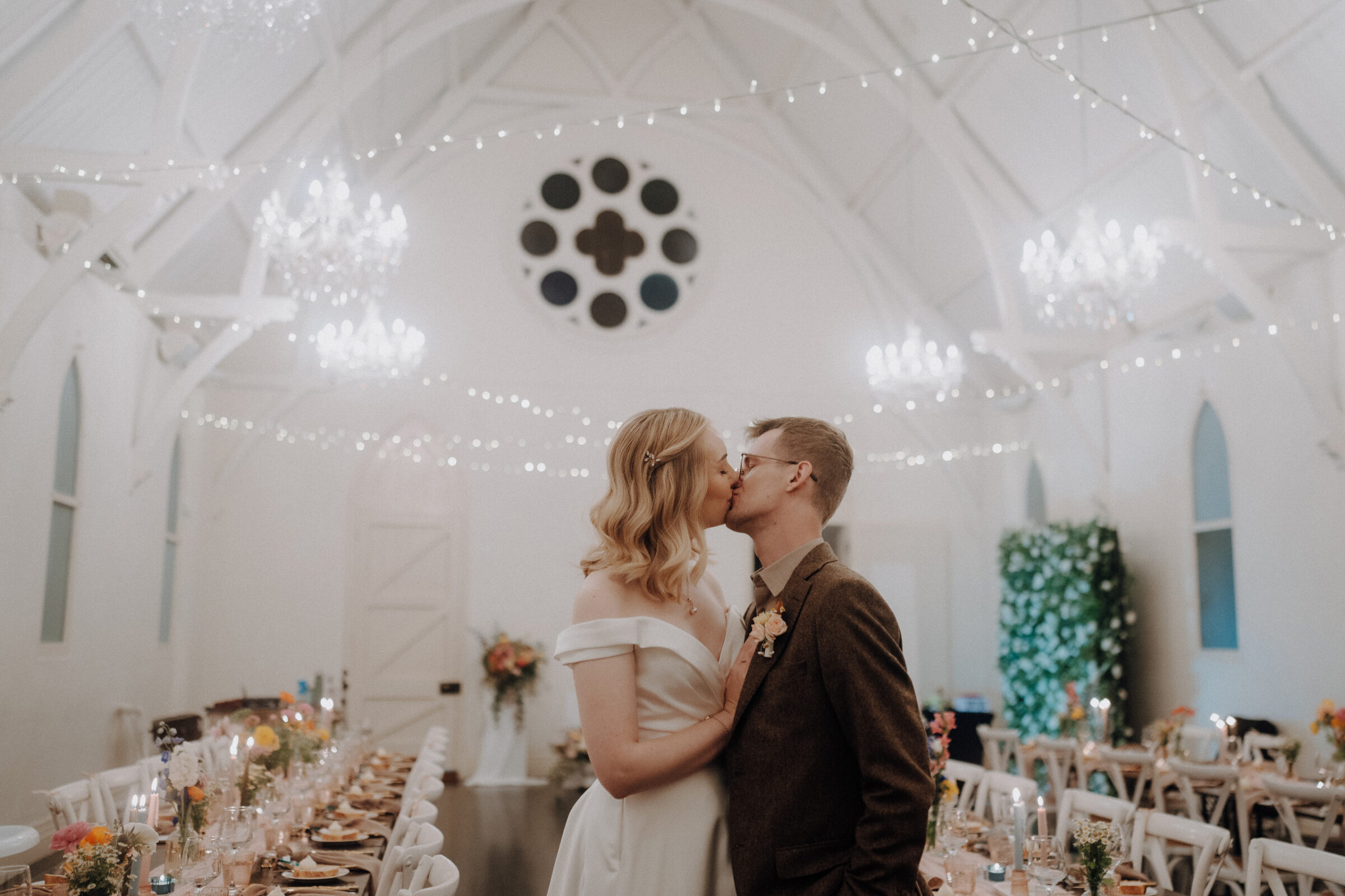 A couple kisses inside a decorated wedding venue with chandeliers, string lights, and a long banquet table.