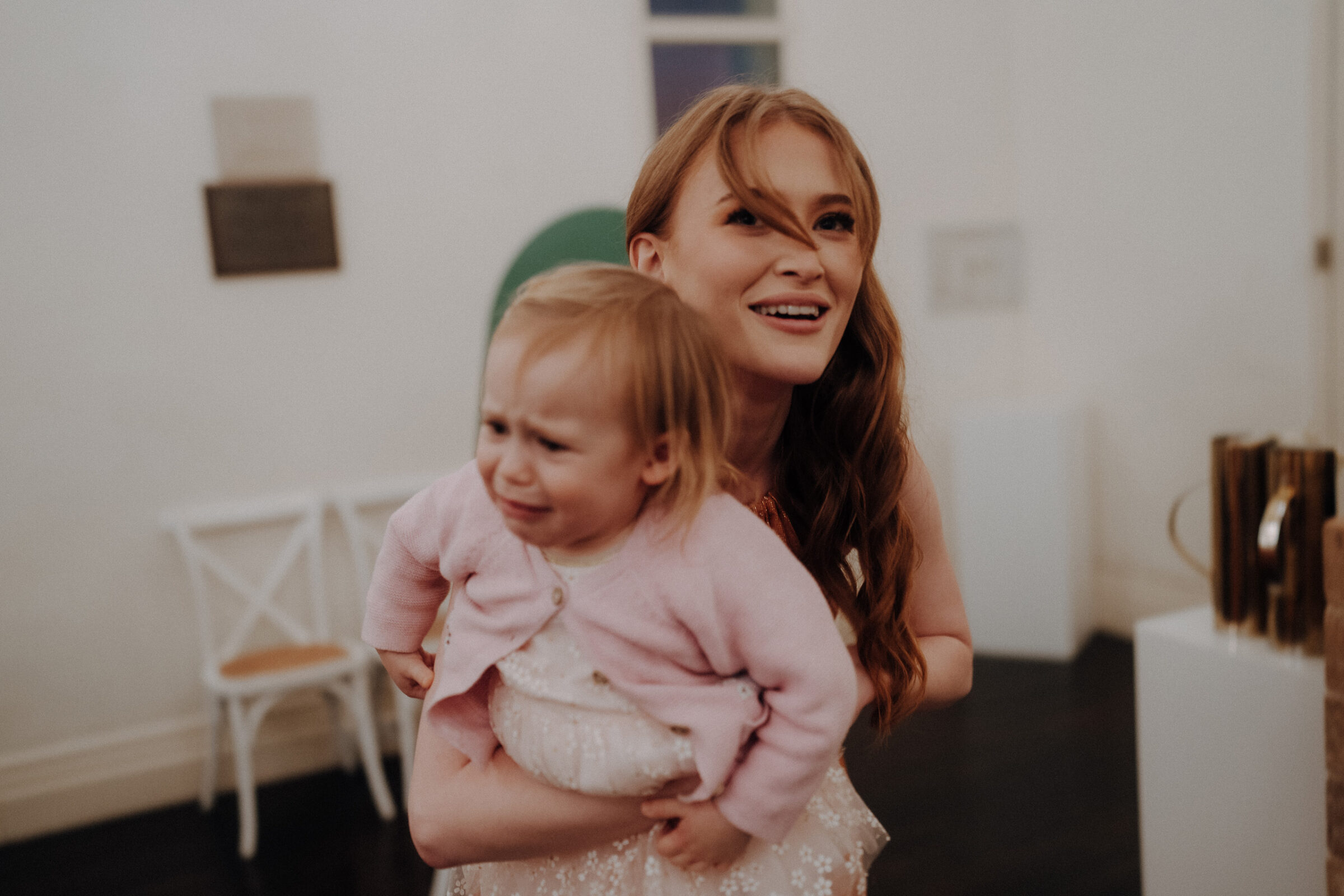 A woman smiles while holding a crying toddler in a white room with minimal decor.