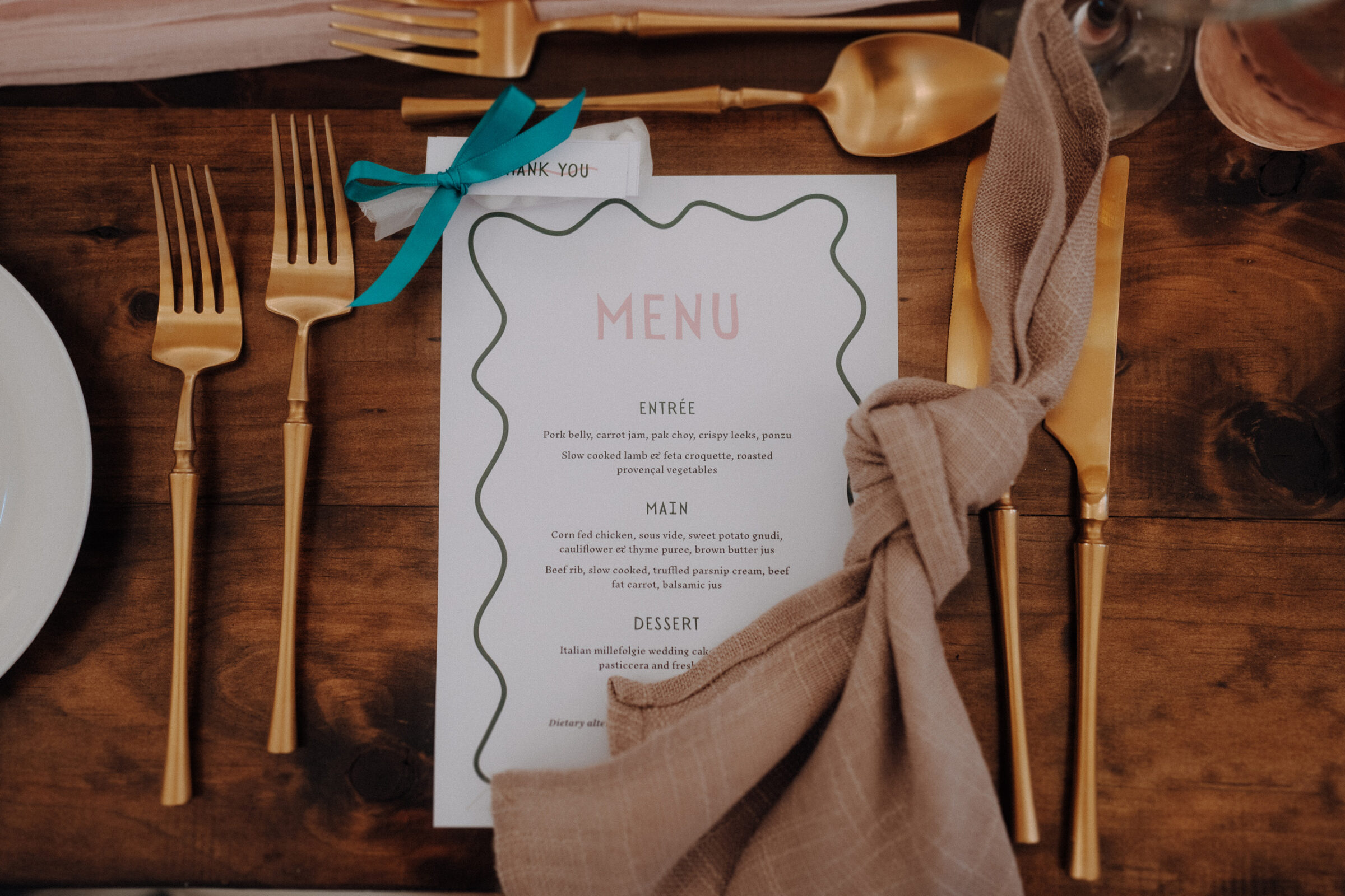 A menu on a wooden table is flanked by gold cutlery and a tan napkin. A blue ribbon is tied around the top left corner of the menu.