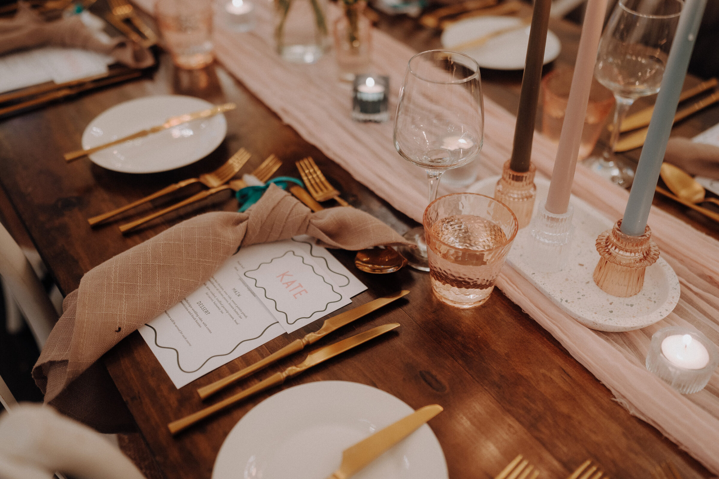 A wooden table set for a formal dinner, featuring plates, gold cutlery, a menu, a folded napkin, glasses, and tall candles on a pink runner.