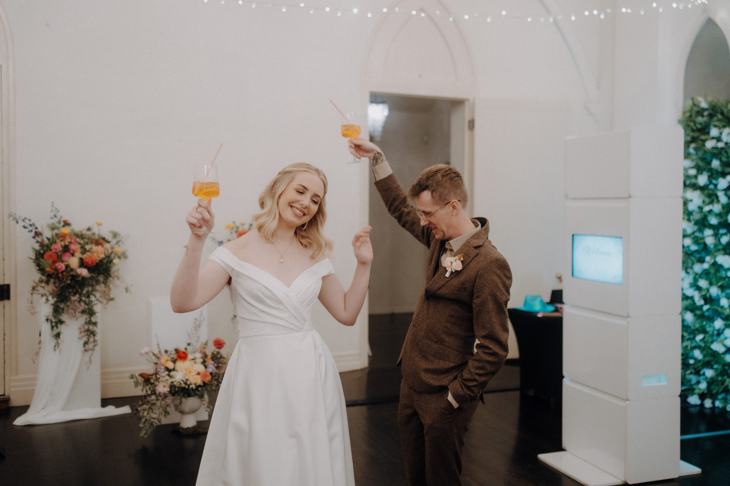 A bride in a white dress and a groom in a brown suit hold drinks and raise their arms while dancing in a decorated room.