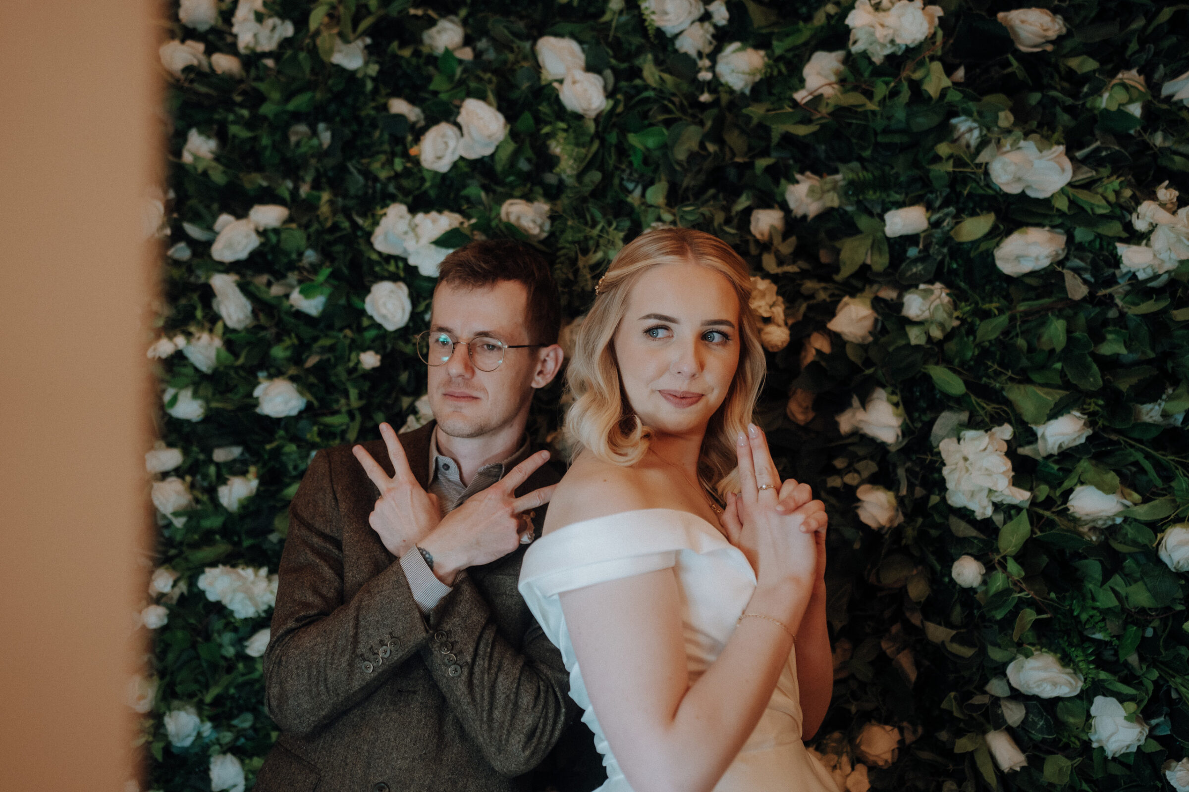 A man and woman pose playfully back-to-back, mimicking finger guns, in front of a backdrop of white roses and greenery.