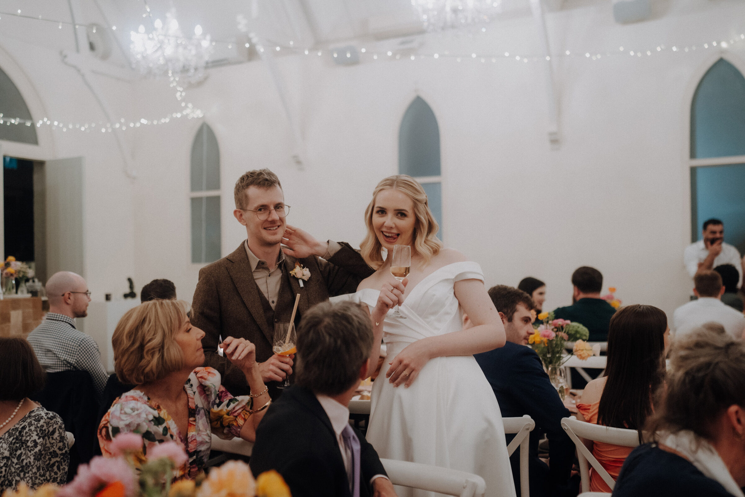 A couple, including a man in a brown suit and a woman in a white dress, stands smiling at a wedding reception. Guests are seated at tables decorated with flowers.