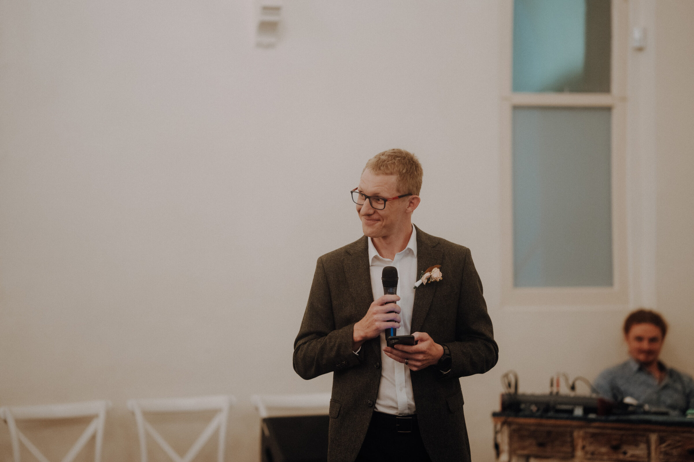 A person in a suit holds a microphone while standing near a white wall, with a seated individual and audio equipment in the background.