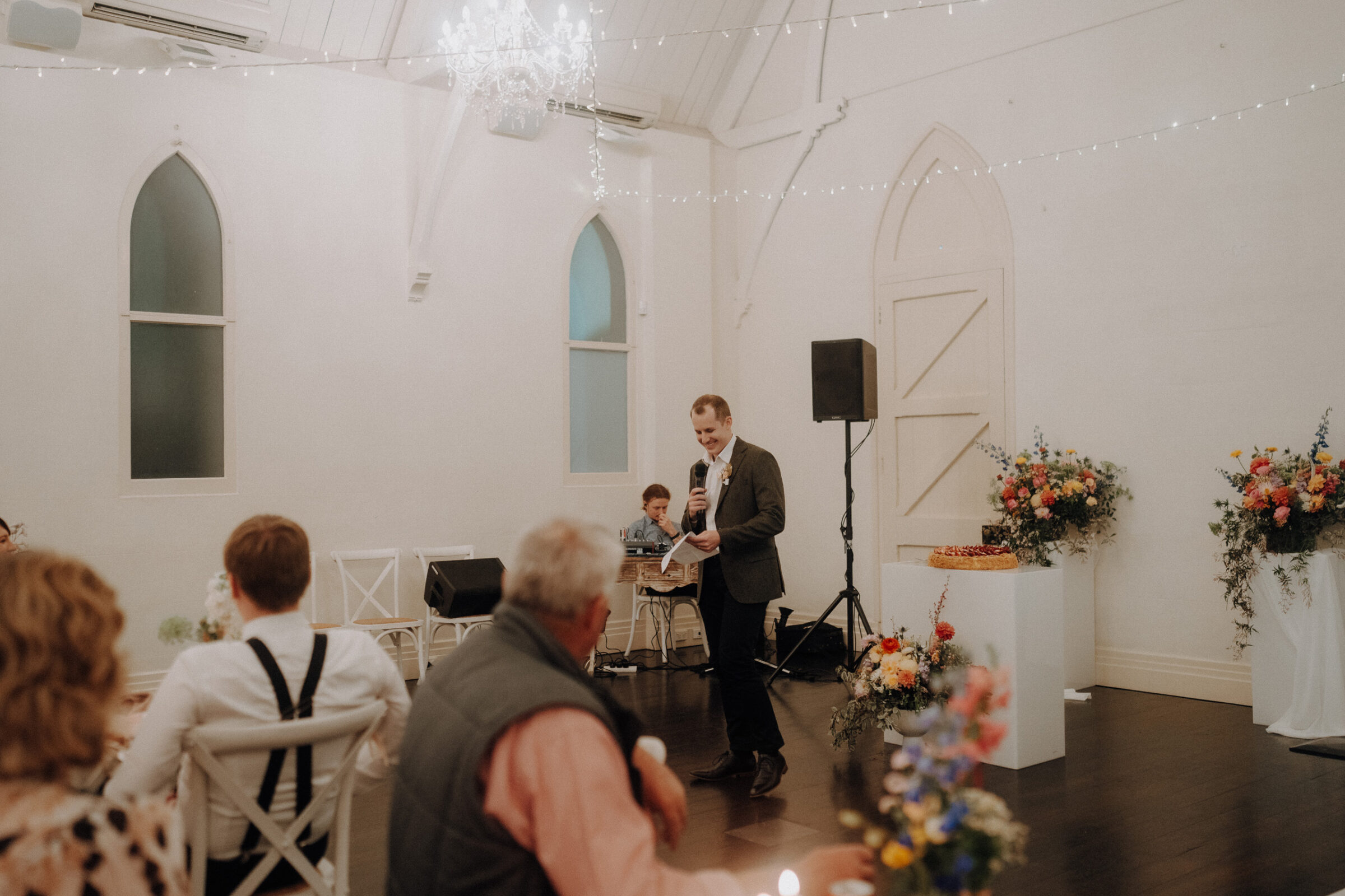 A man in a suit gives a speech in a decorated room with an audience seated. There are floral arrangements and a white chandelier.