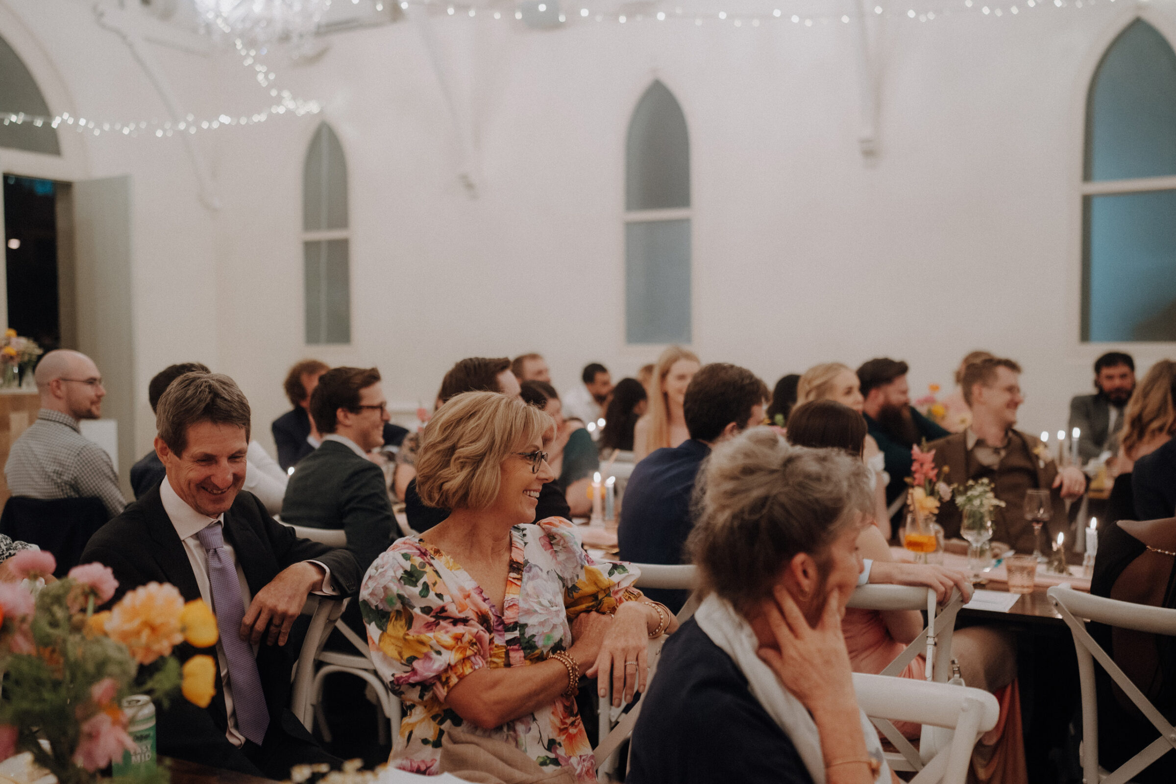 People seated at tables in a softly lit, decorated room, with flowers and candles, attending a social gathering or event.