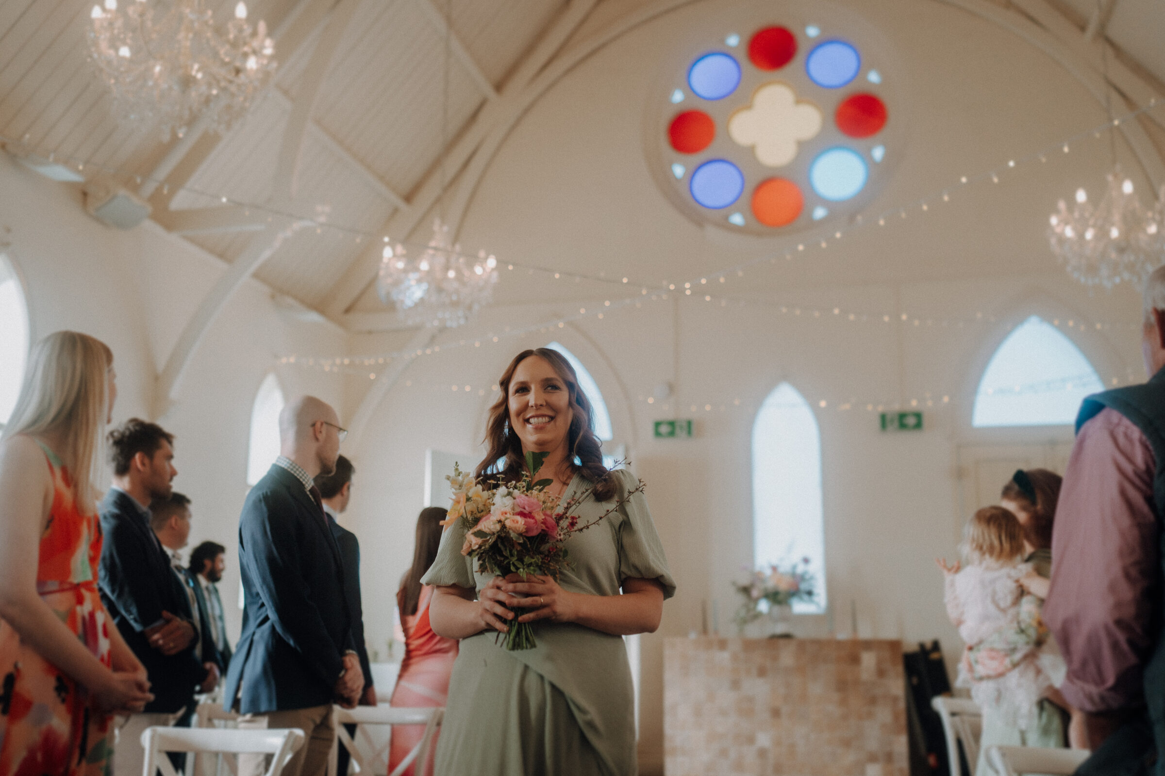 A woman in a green dress holds a bouquet in a chapel with a colorful stained-glass window and string lights above. People are seated around her.