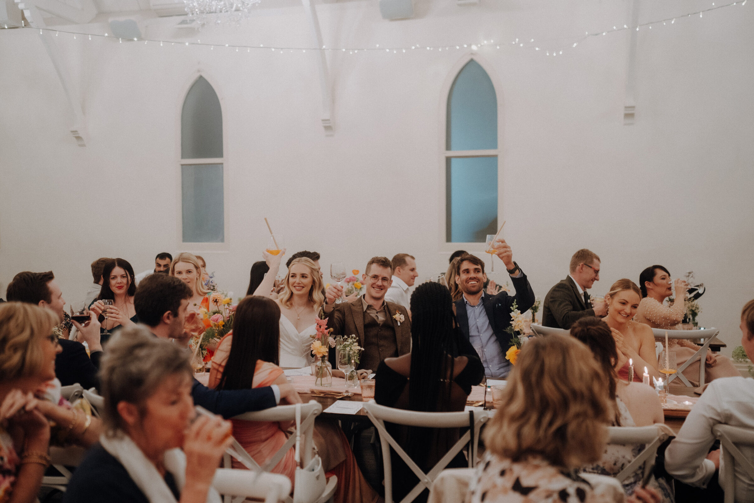 Guests seated around a long table at a wedding reception, raising glasses in a toast. The room is decorated with flowers and string lights.