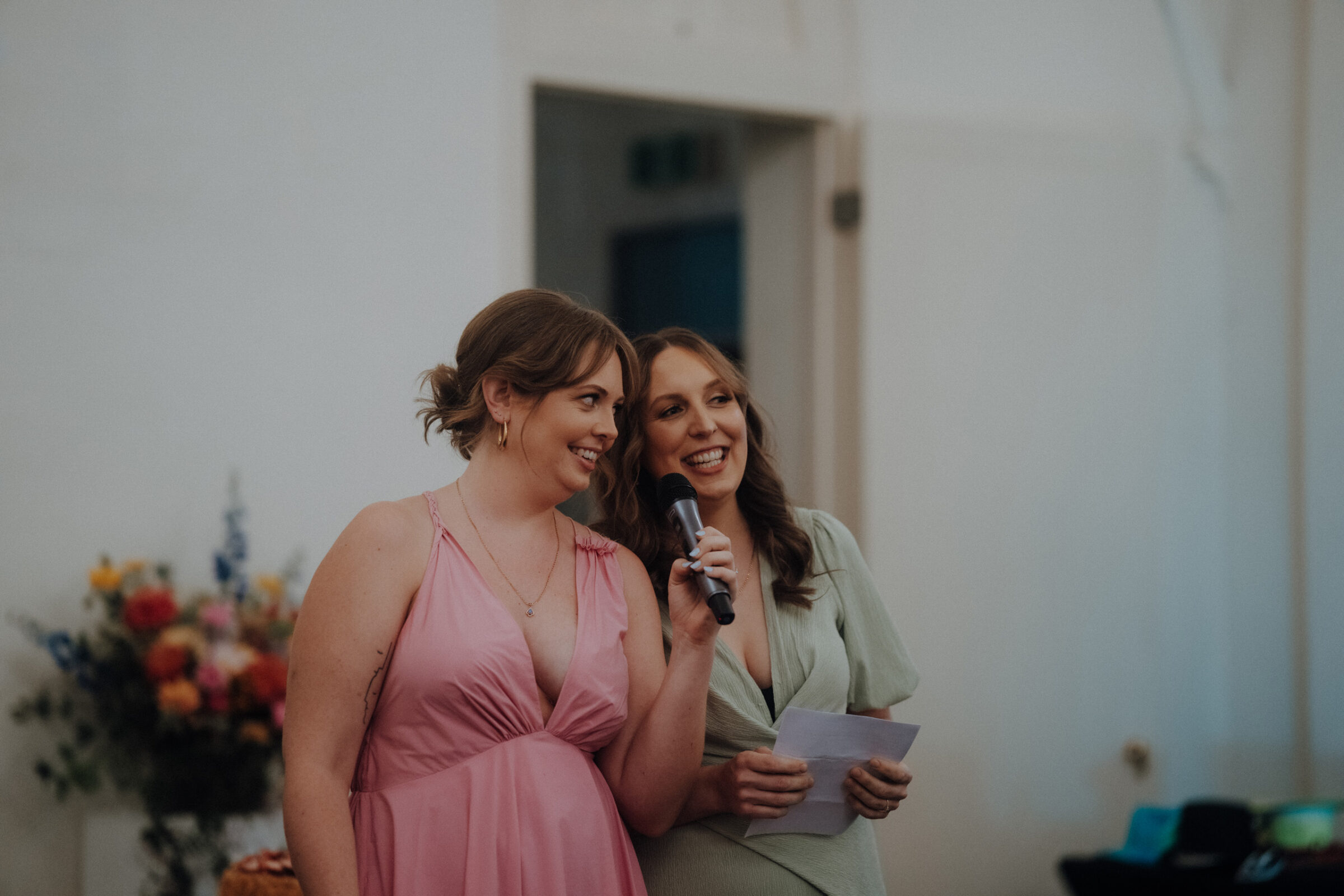 Two women in pastel dresses stand together. One holds a microphone, the other a piece of paper. Flowers are visible in the background.