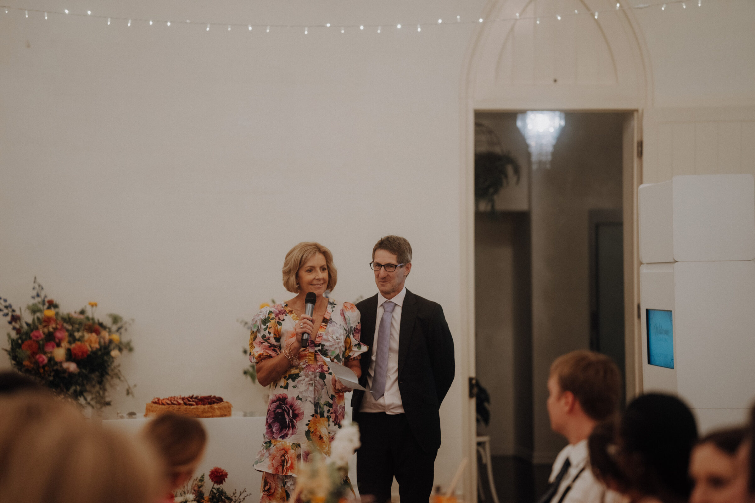 A woman in a floral dress speaks into a microphone beside a man in a suit at a wedding reception.