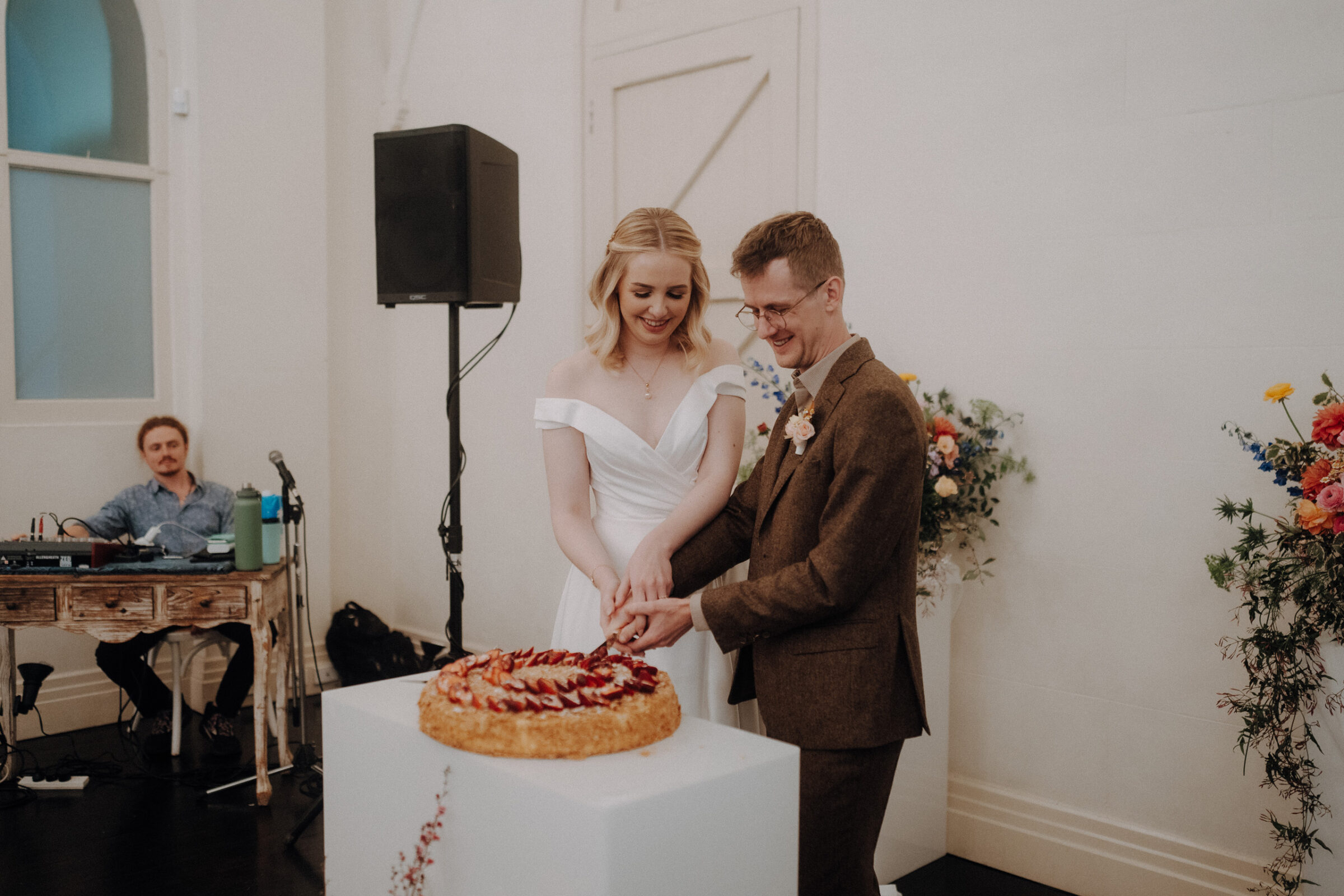 A couple in wedding attire smiles while cutting a large pie on a white pedestal. A person in the background is seated at a desk with sound equipment.