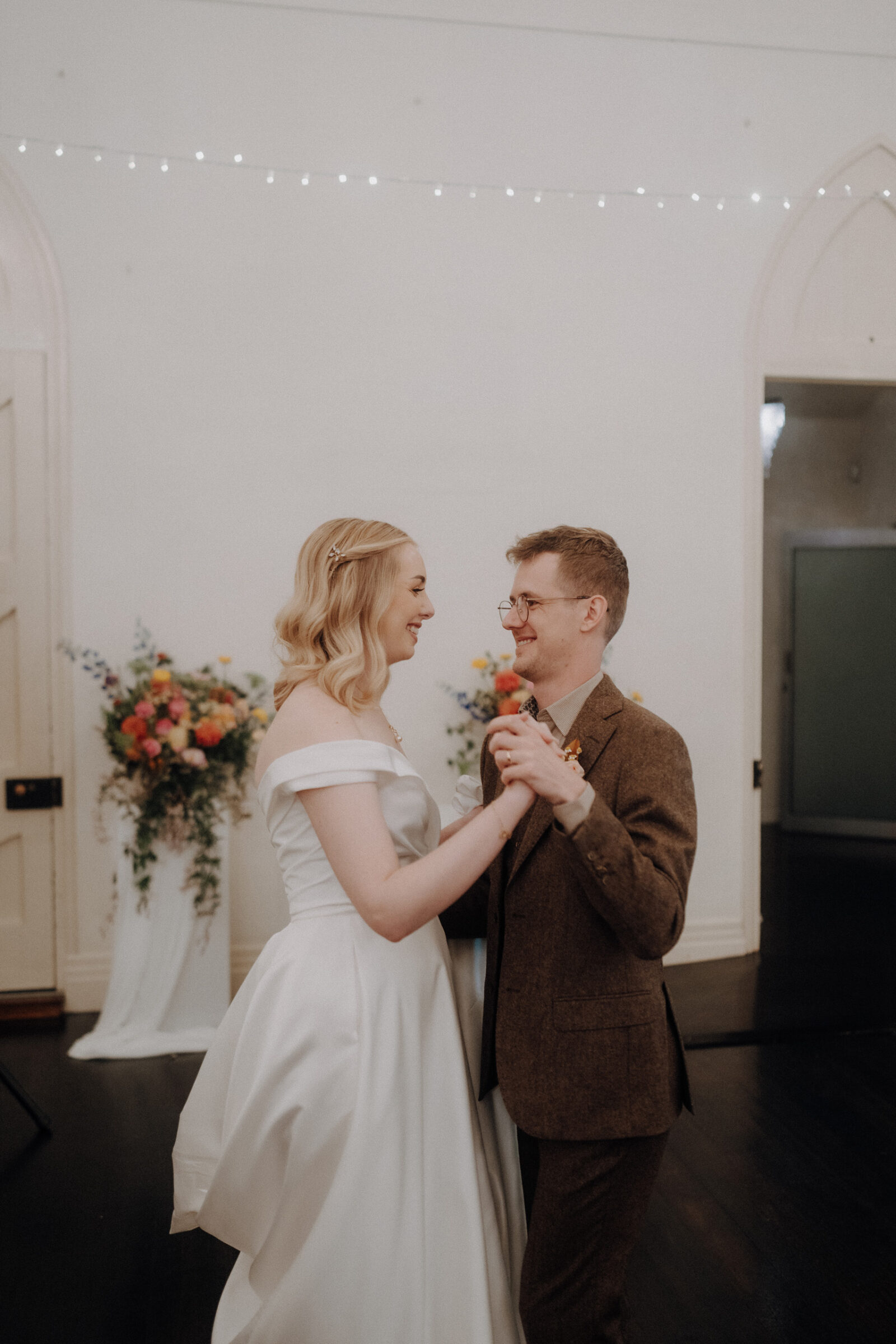 A couple dances in an indoor setting with flowers and string lights in the background. The woman wears a white dress, and the man wears a brown suit.