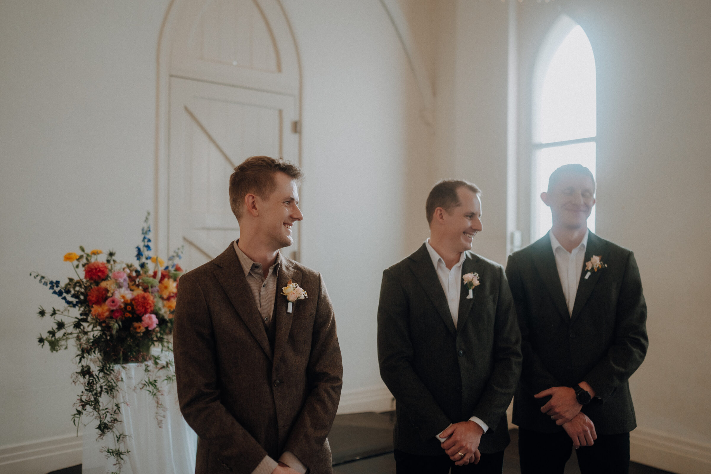 Three men in suits stand in a bright room beside a flower arrangement, smiling and looking to the side.