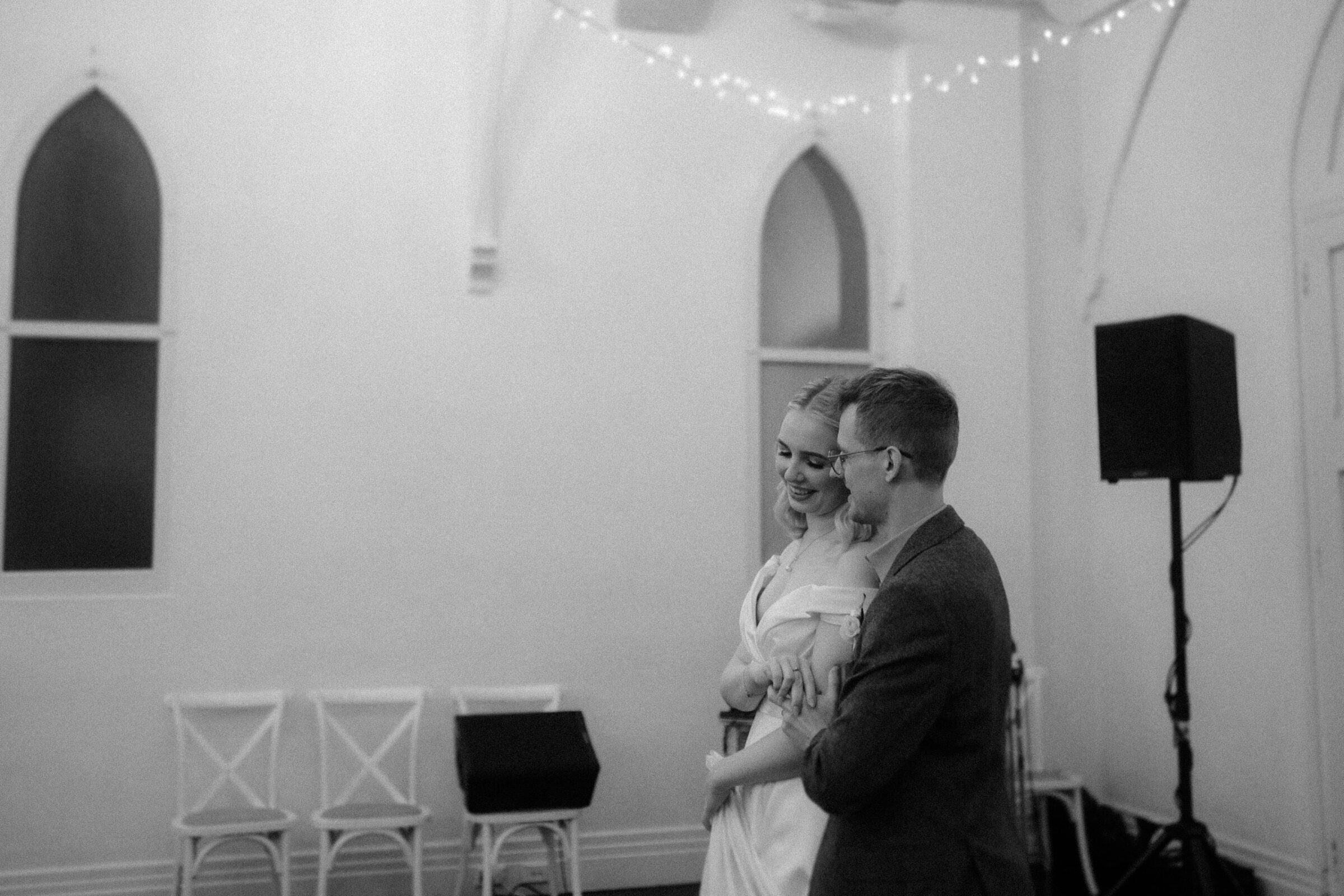 A couple in formal attire dances in a softly lit room with arched windows and string lights.