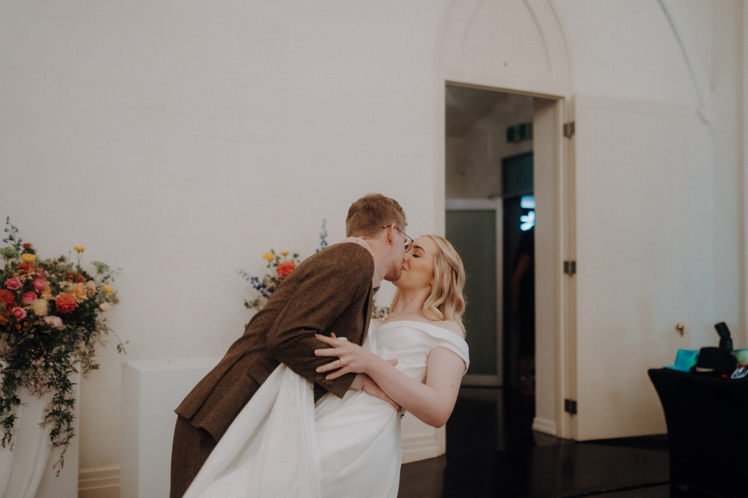 A couple kissing indoors, near a doorway, with floral decorations on the left.