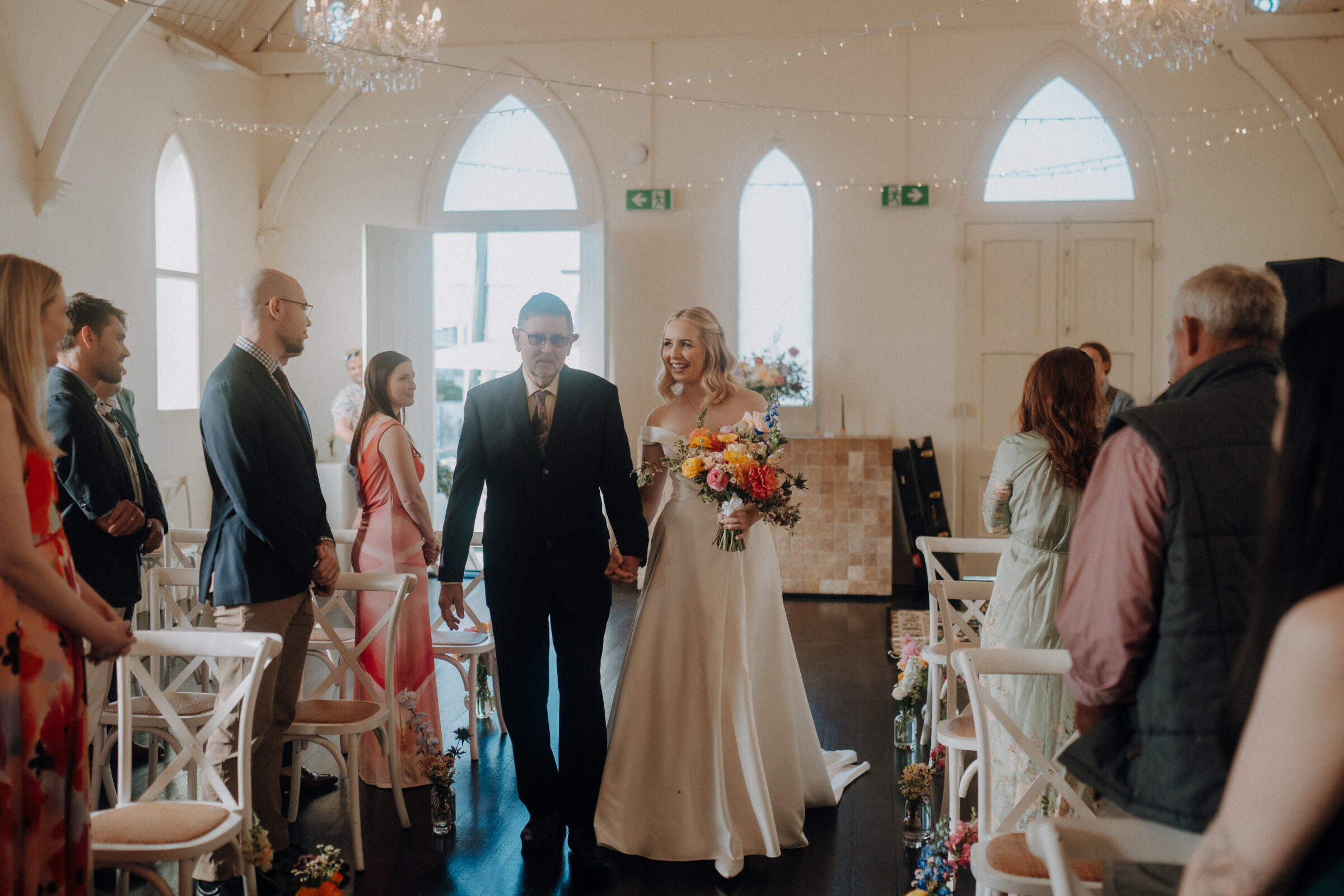 A bride holding flowers walks down the aisle with a man in a suit, surrounded by standing guests in a decorated venue.