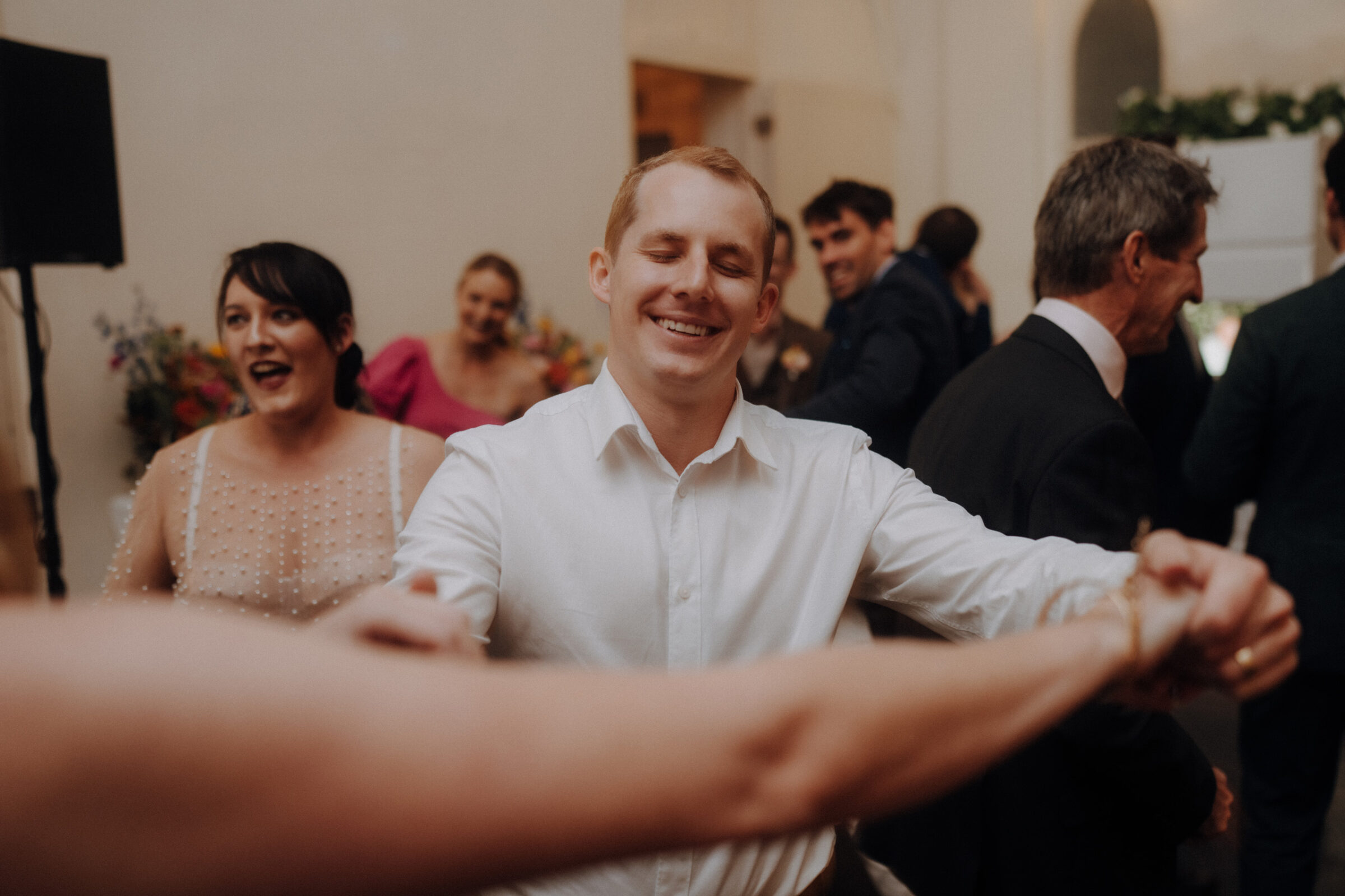 A smiling man in a white shirt dances with a partner at a social event, surrounded by other guests.