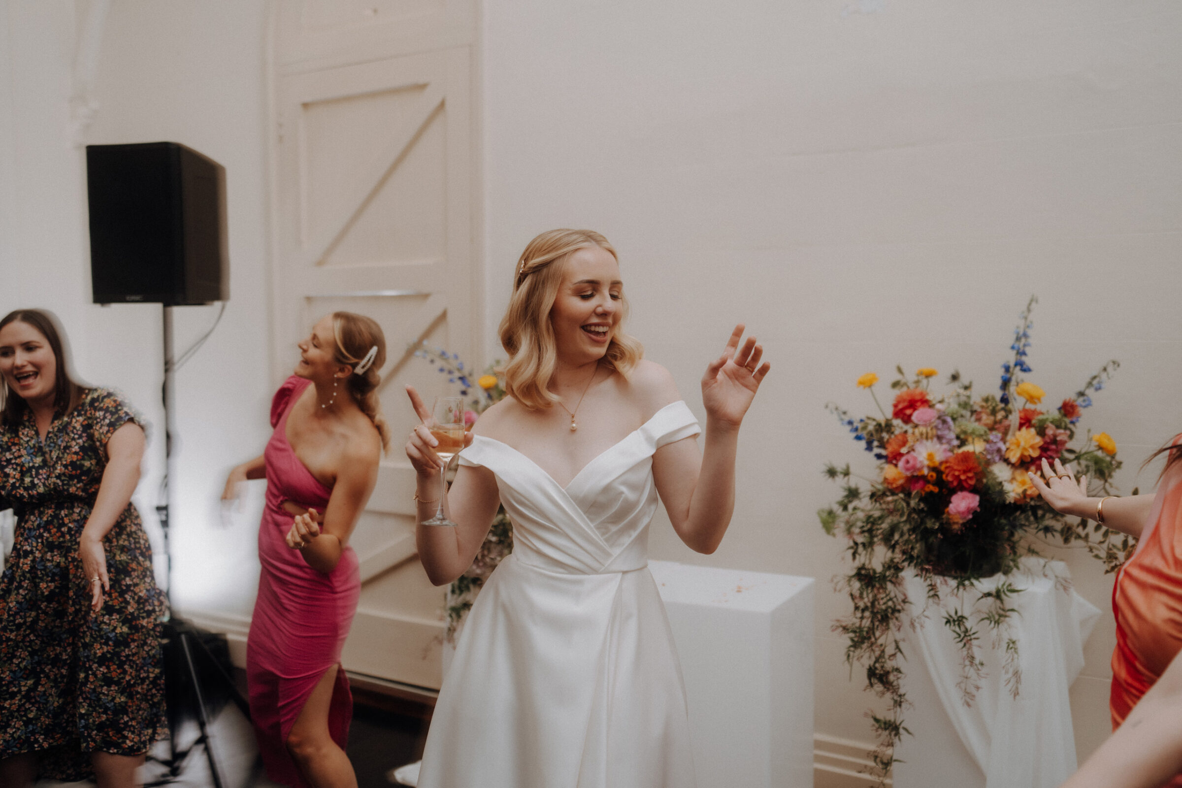 A bride in a white dress smiles and dances with a drink in hand. Three women dance beside her in a room with a floral arrangement in the background.