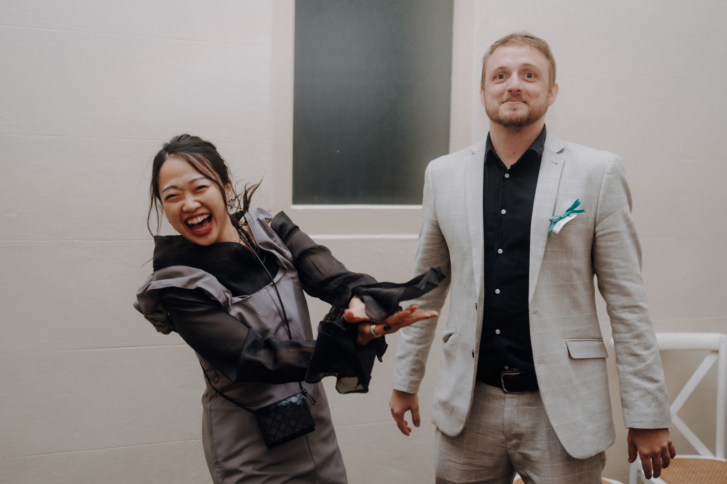 A woman in a black and gray outfit smiles while gesturing beside a man in a light gray suit with a black shirt and teal pocket square. They stand indoors near a white wall and window.