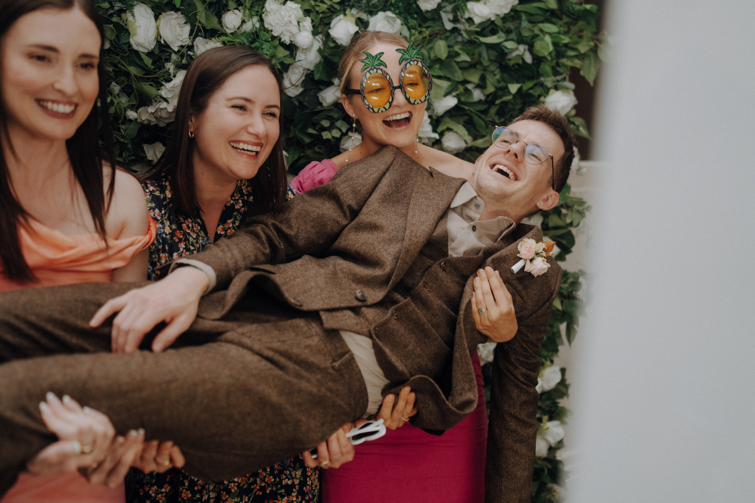 A group of four people laughing, with three women holding up a man in a brown suit. One woman wears pineapple-shaped glasses. A leafy backdrop is behind them.