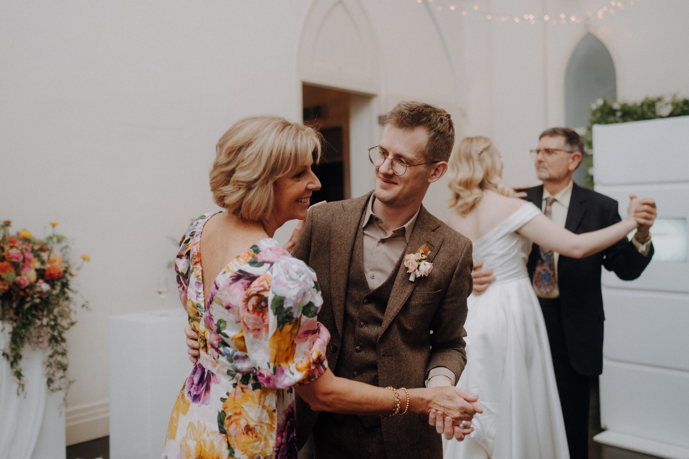 Two pairs of people are dancing indoors. A man in a brown suit dances with a woman in a floral dress. Another couple is in the background. There are flowers on the left.