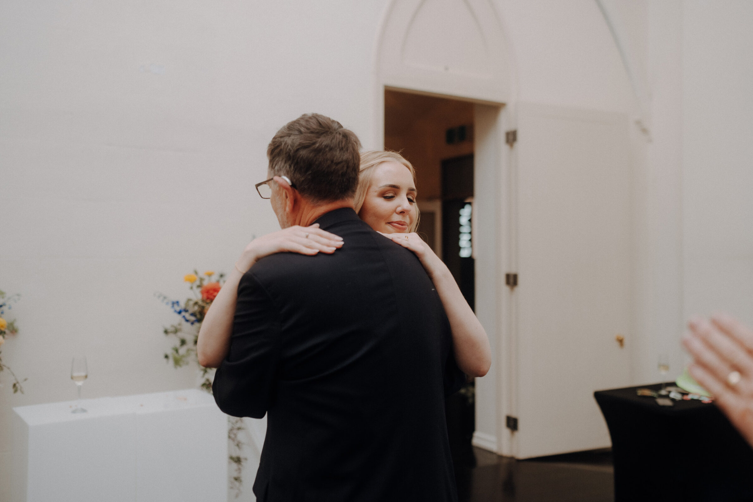 A couple embraces in a warmly lit room, surrounded by minimal floral decorations.