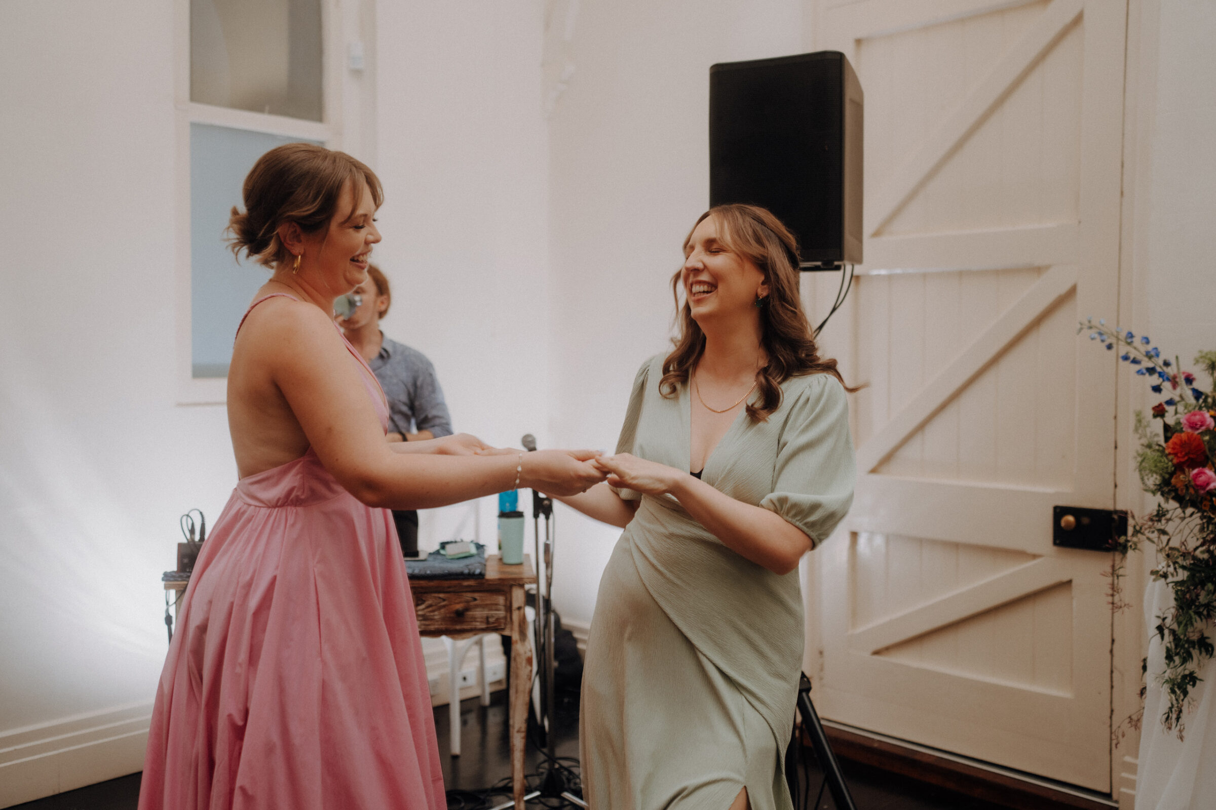 Two women in dresses dance together in a room with a wooden door and a speaker in the background. Flowers are visible on the right side.