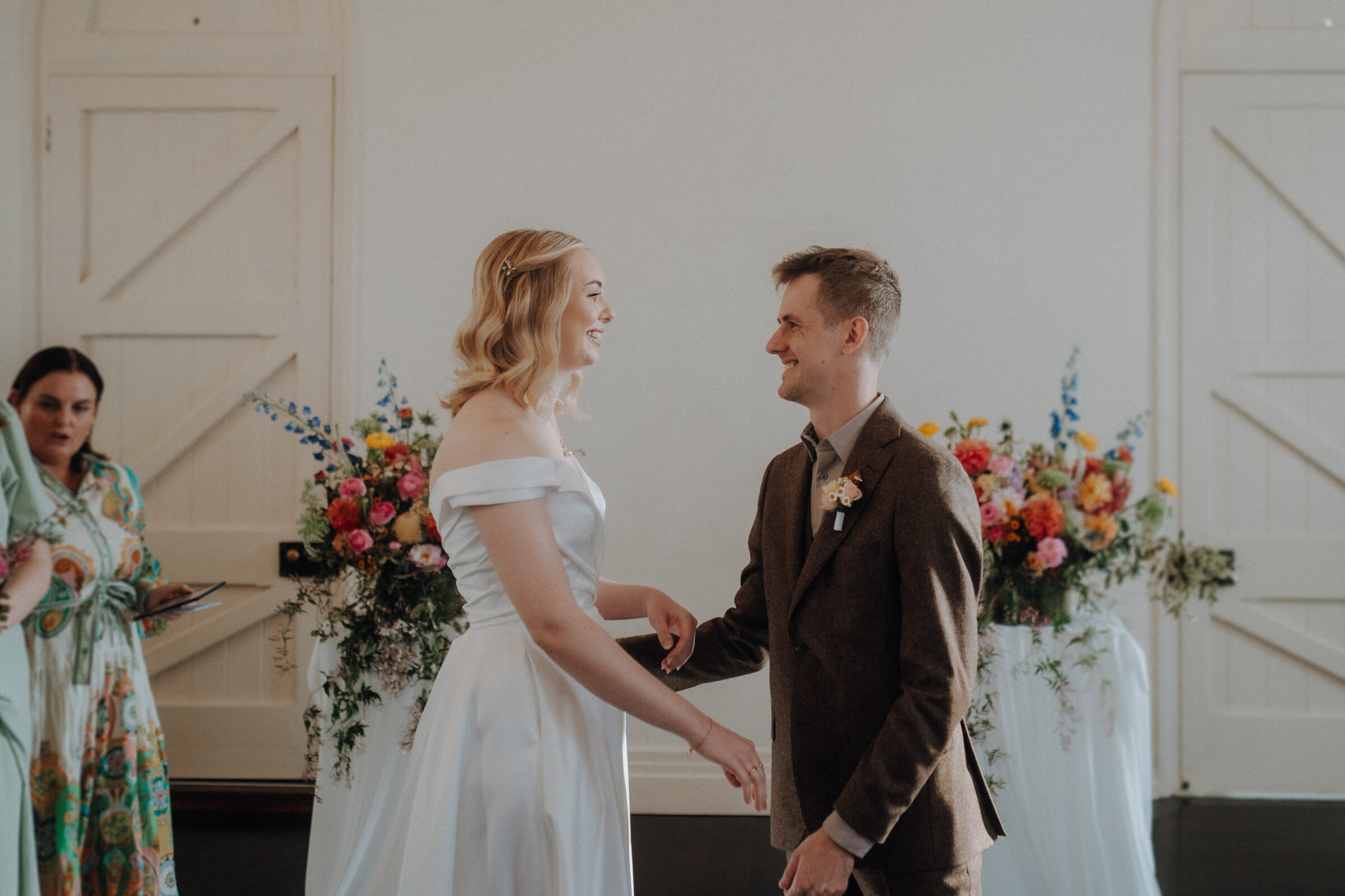 A bride and groom smile at each other during their wedding ceremony, with a woman in the background and colorful floral arrangements behind them.