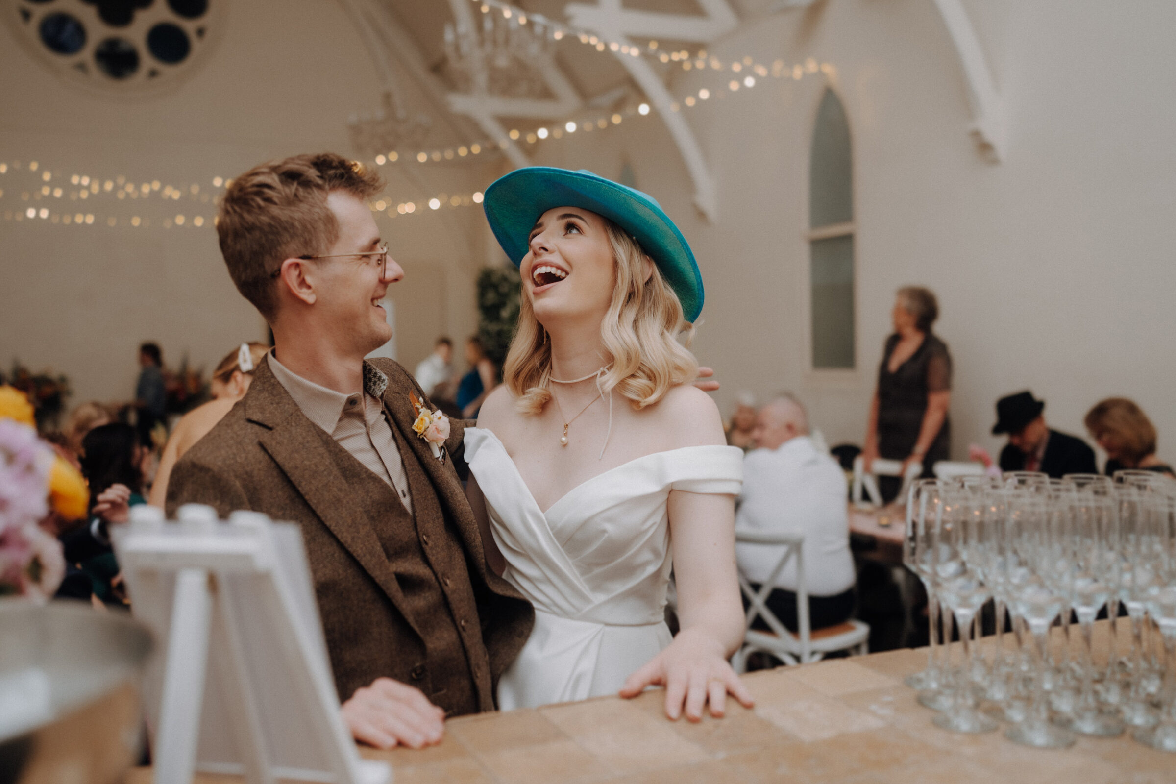 A bride and groom in a lively venue share a joyful moment. She wears a white dress and blue hat, while he is in a brown suit. Champagne glasses are on the counter beside them.