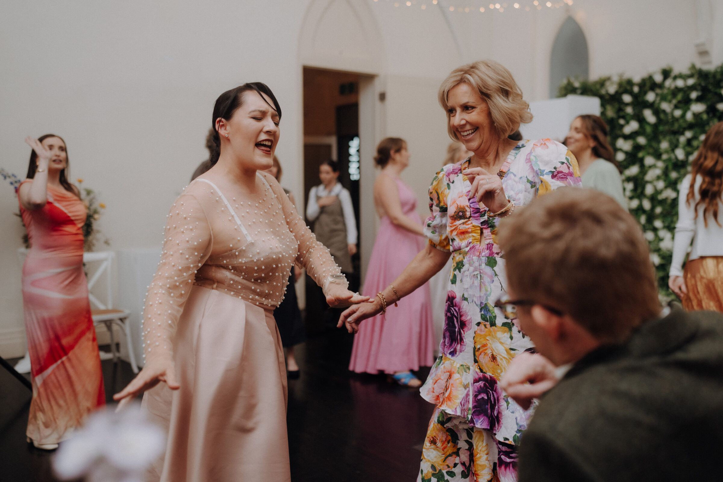 People dancing and smiling at a celebration, with one person in a pink dress and another in a floral dress, surrounded by others in colorful attire.