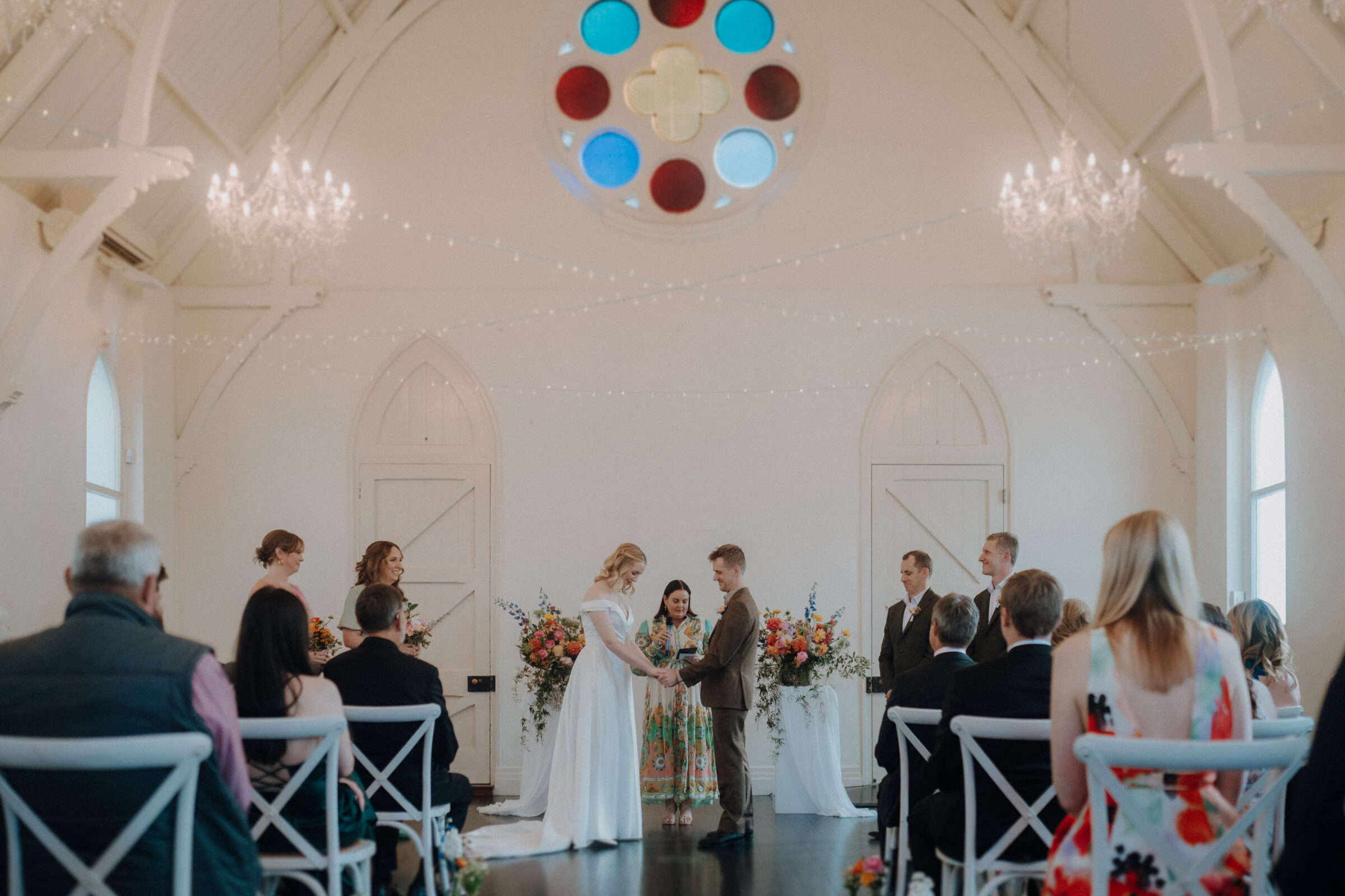 A couple stands facing each other in a bright, decorated chapel with guests seated. A celebrant stands between them. Chandeliers and a round stained glass window are visible.