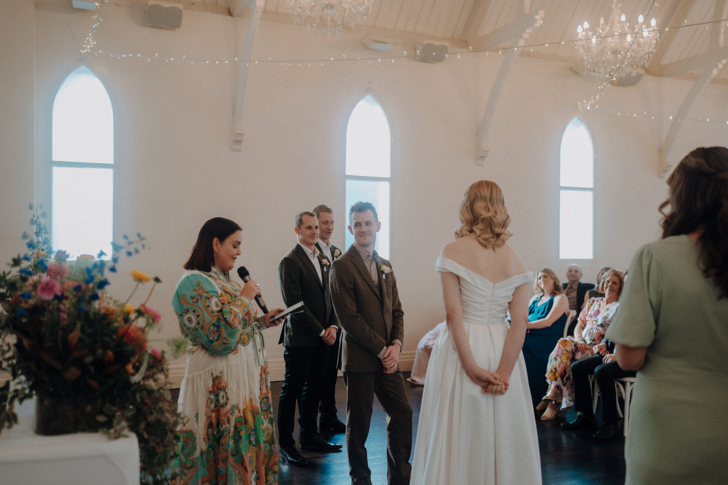 A wedding ceremony in a bright chapel with a bride in a white gown and a groom in a suit. A woman is speaking into a microphone, and guests are seated nearby. Flowers and chandeliers decorate the space.