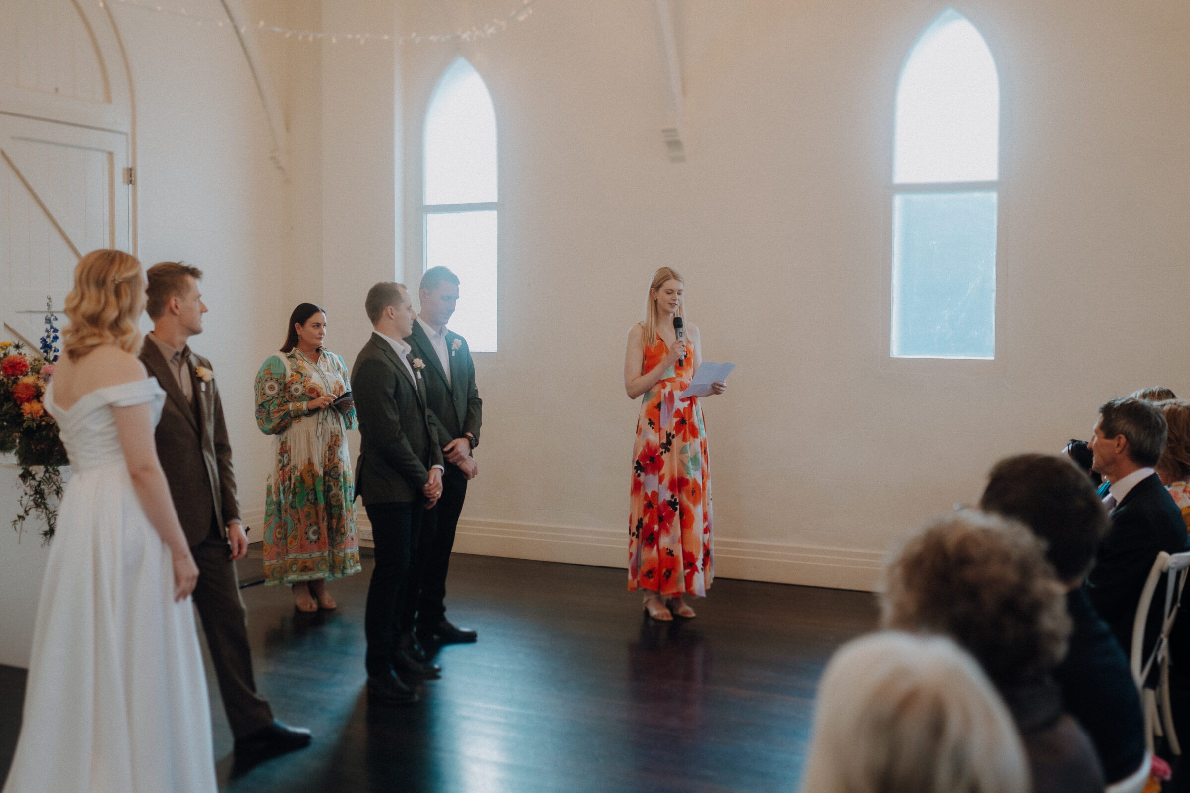 A person in a colorful dress speaks into a microphone at a wedding ceremony, with the couple and others standing nearby in a brightly lit room.