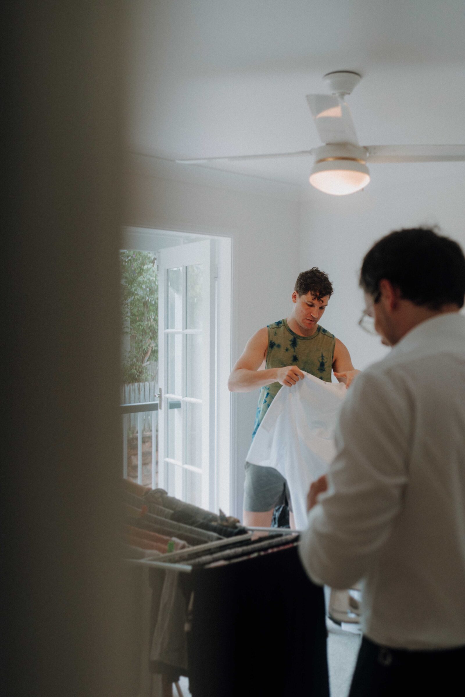 Two people in a room doing laundry, one holds a white shirt, and the other stands near a rack with clothes. A ceiling fan is above, and sunlight enters from an open door.